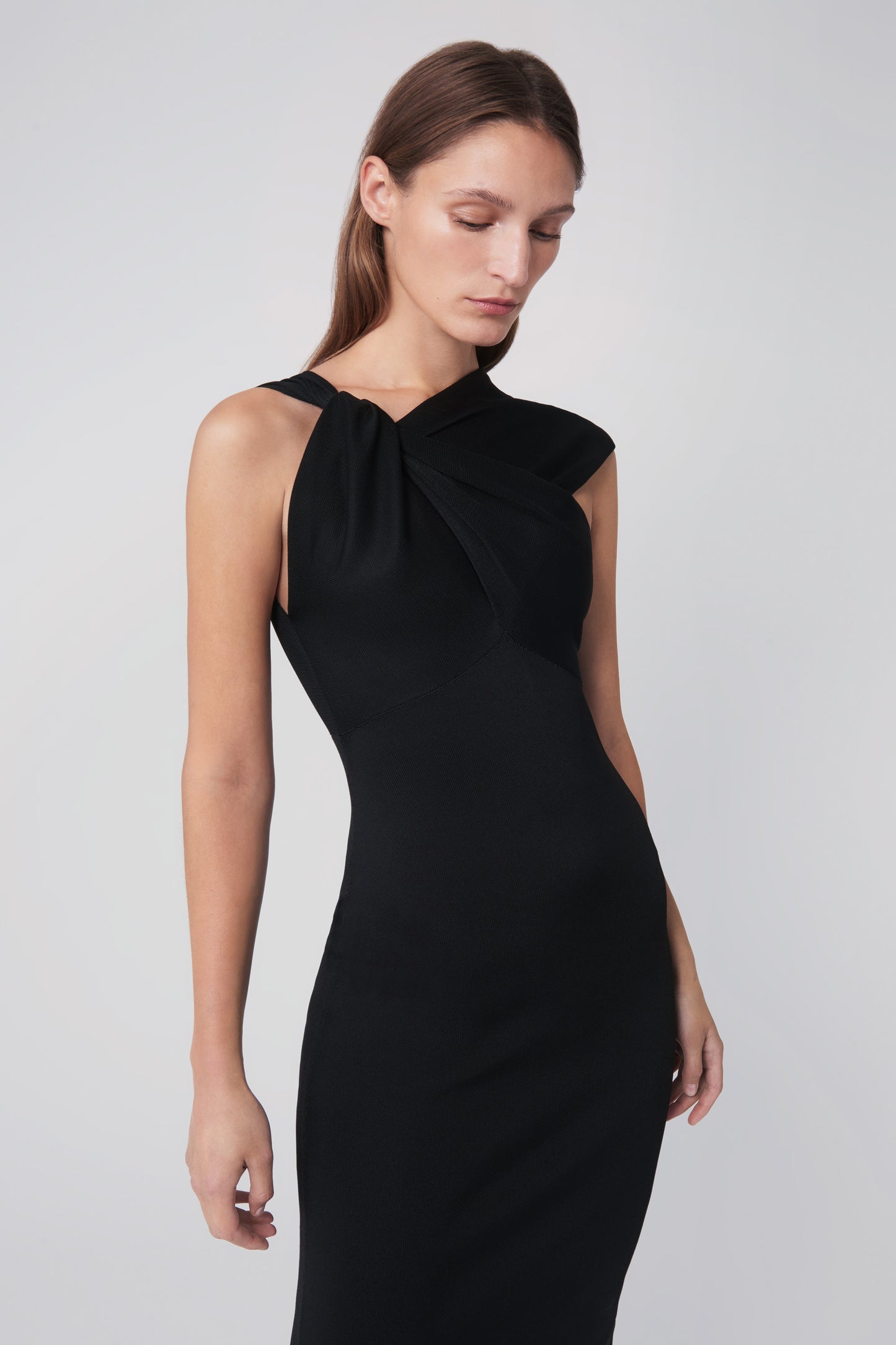 A woman wearing the Twist Drape Knit Dress in Black by Victoria Beckham, featuring crossed straps and an asymmetric neckline, looks downward against a plain background.