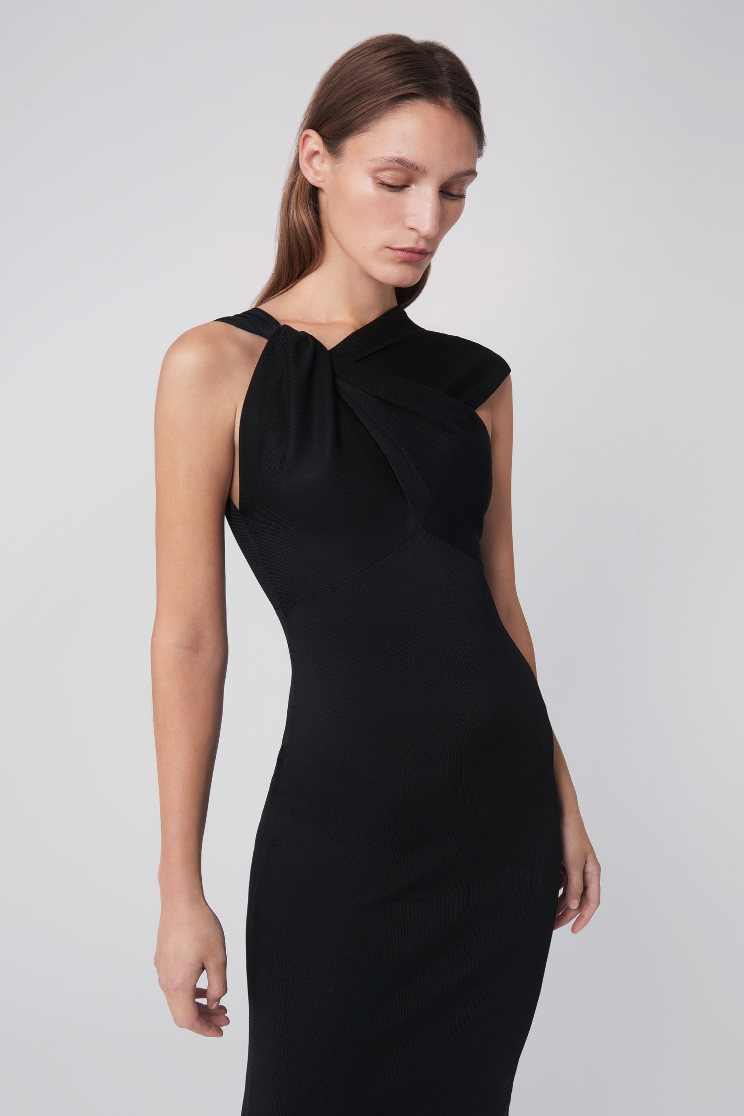 A woman wearing the Twist Drape Knit Dress in Black by Victoria Beckham, featuring crossed straps and an asymmetric neckline, looks downward against a plain background.