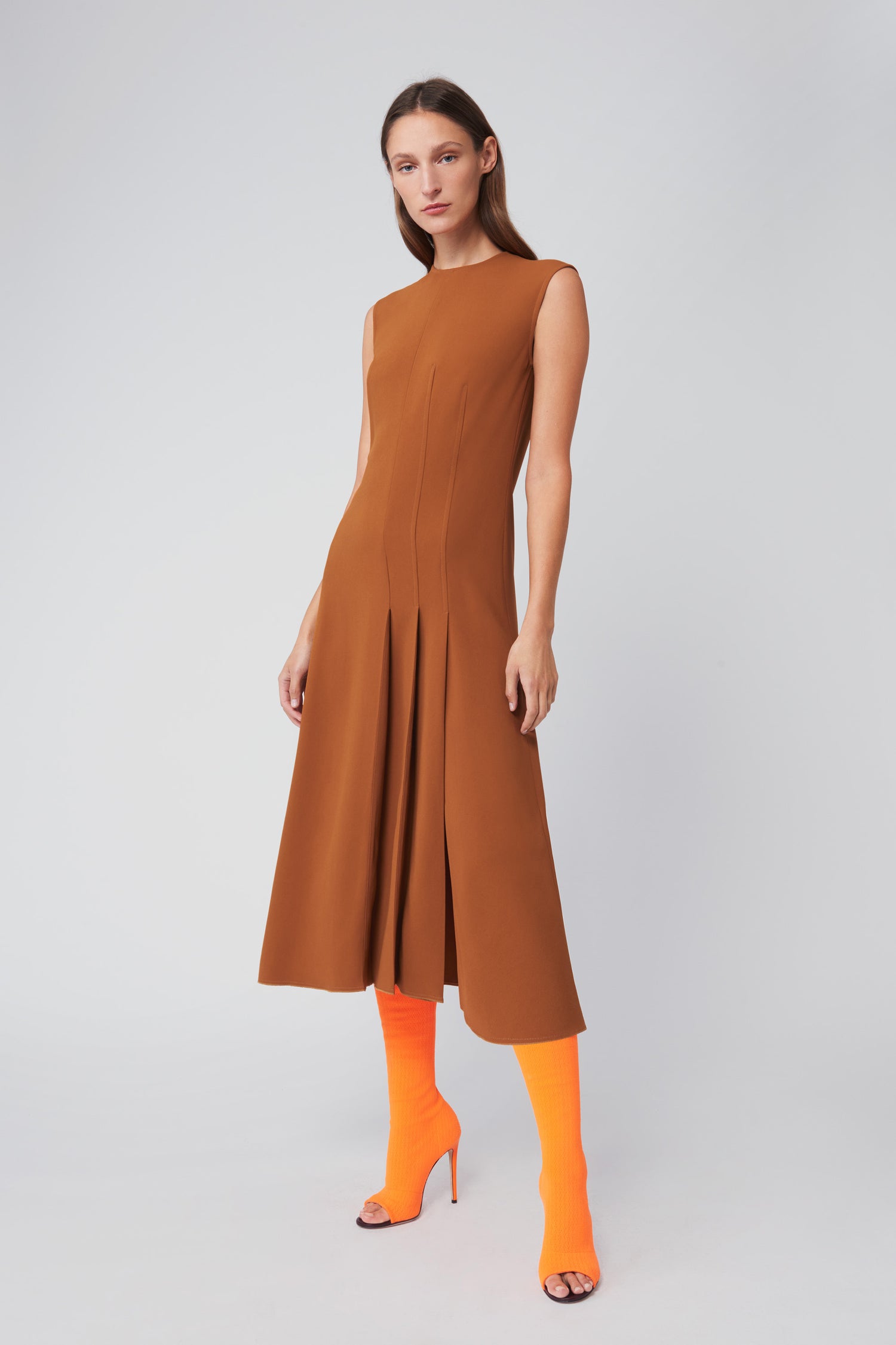 Dressed in a caramel Asymmetric Pleat Drape Dress by Victoria Beckham, an individual pairs it with bright orange knee-high socks and open-toed heels, standing against a plain background.