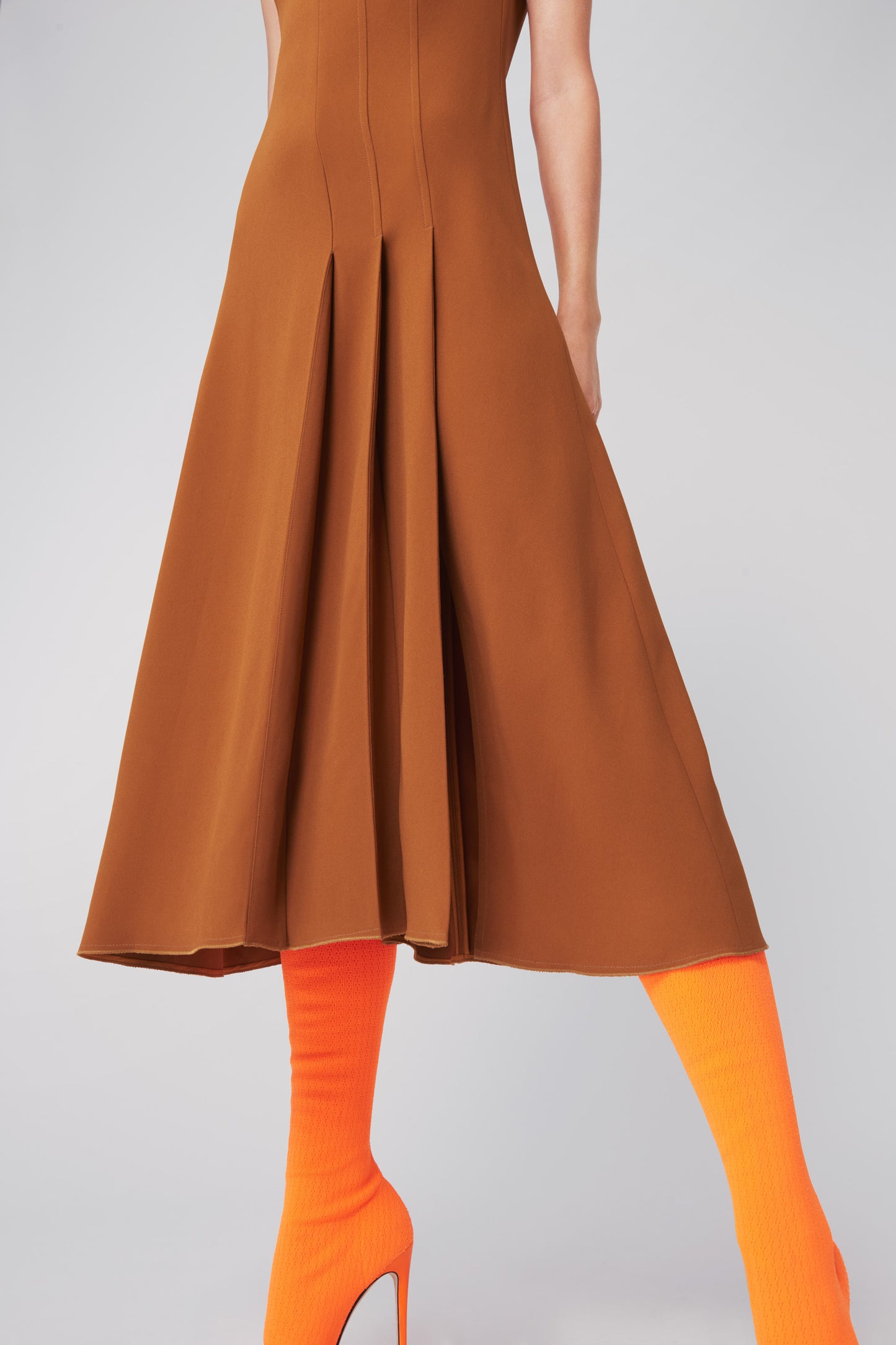 Wearing Victoria Beckham's Asymmetric Pleat Drape Dress in Caramel, paired with bright orange tights and heels, the model poses with one leg forward, highlighting a sinuous silhouette.
