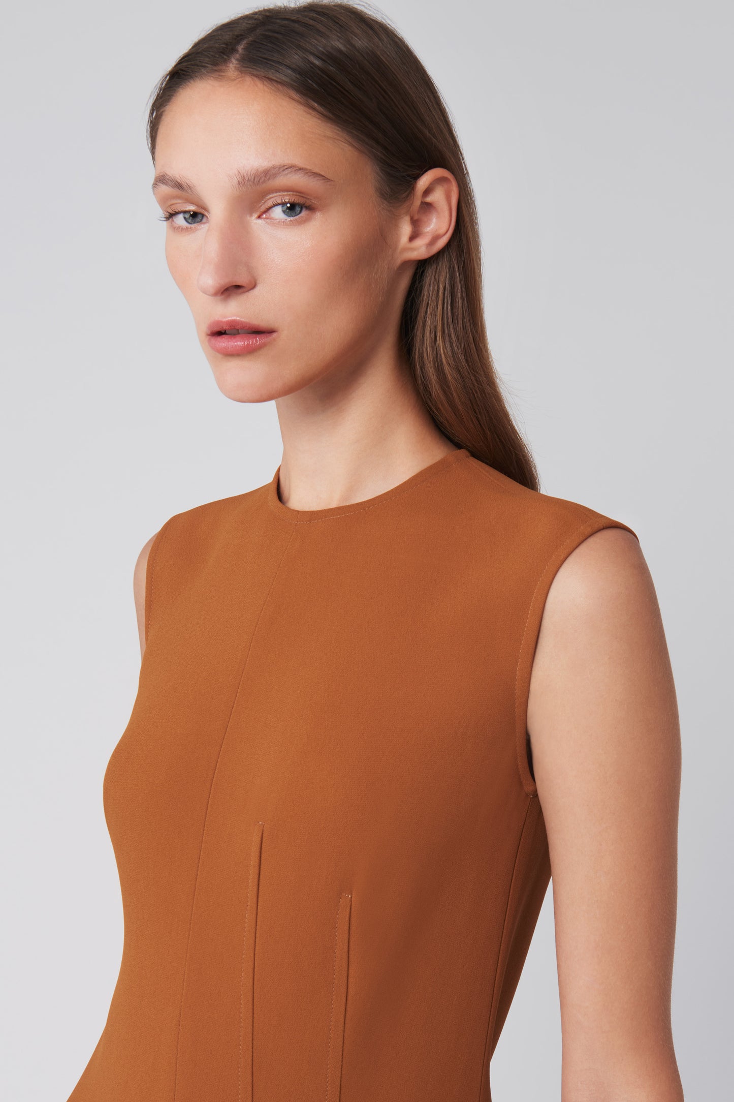 A person wears Victoria Beckham's Asymmetric Pleat Drape Dress in Caramel, featuring a sinuous silhouette crafted from fluid cady fabric, as they gaze slightly left against a plain background.