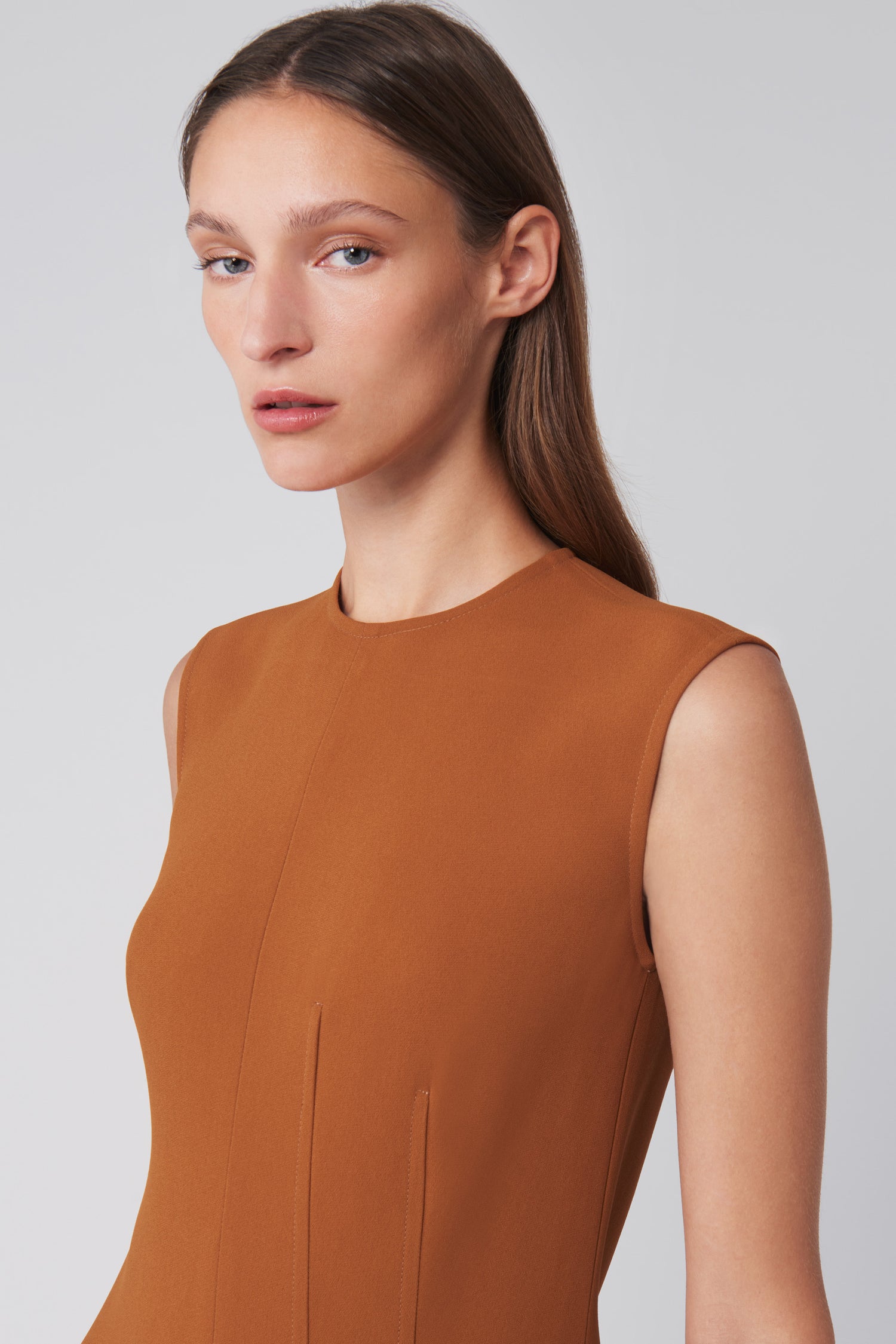 A person wears Victoria Beckham's Asymmetric Pleat Drape Dress in Caramel, featuring a sinuous silhouette crafted from fluid cady fabric, as they gaze slightly left against a plain background.