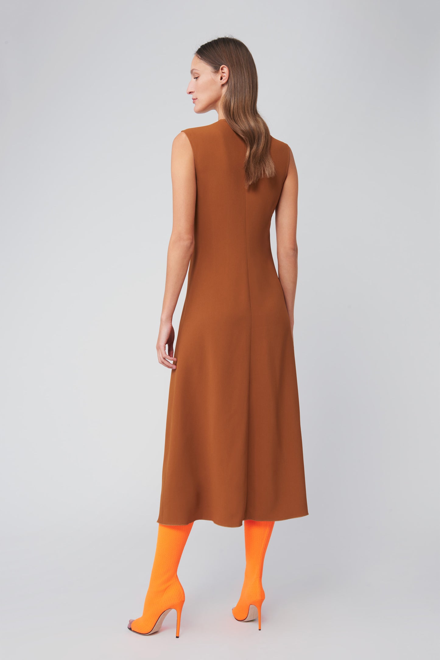 In a plain light setting, a person with long hair poses sideways, highlighting their silhouette in Victoria Beckham's Asymmetric Pleat Drape Dress in caramel, paired with bright orange socks and matching high heels.