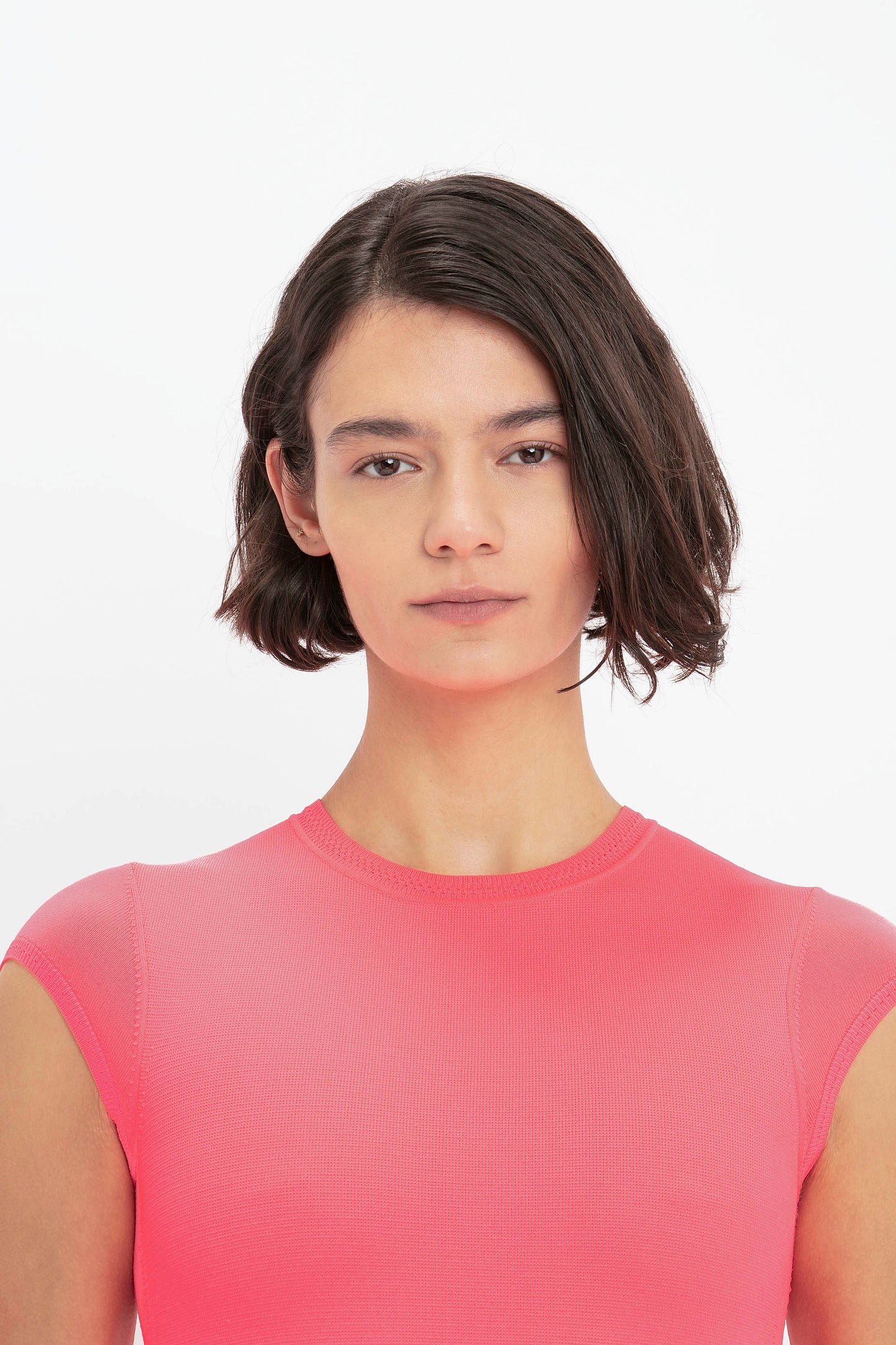 A young woman with short, straight dark hair stands against a plain white background, wearing the VB Body Compact Cap Sleeve Mini Dress in Pink by Victoria Beckham.