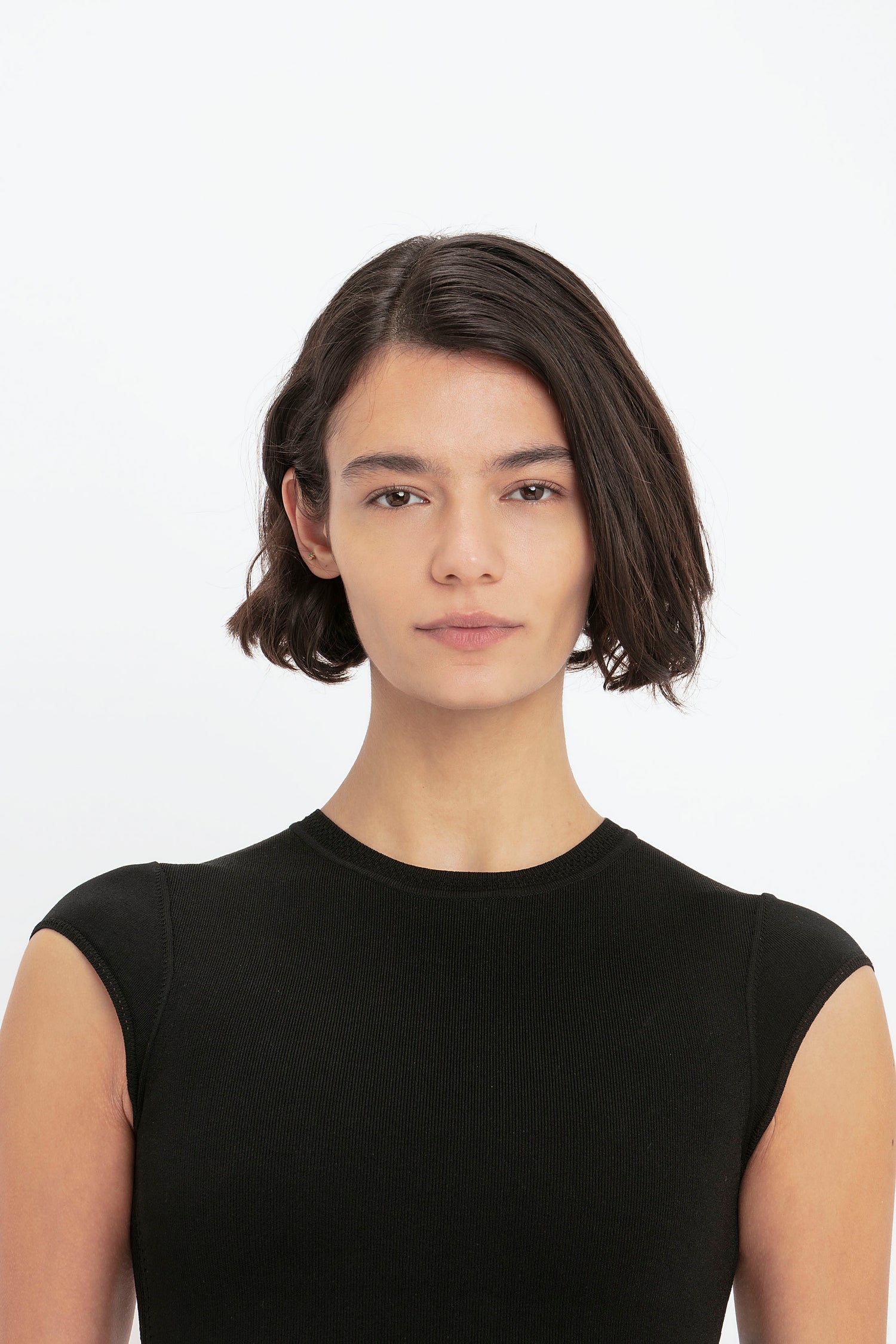 A person with short brown hair and a neutral expression, wearing a VB Body Compact Cap Sleeve Mini Dress in Black by Victoria Beckham, photographed against a plain white background.