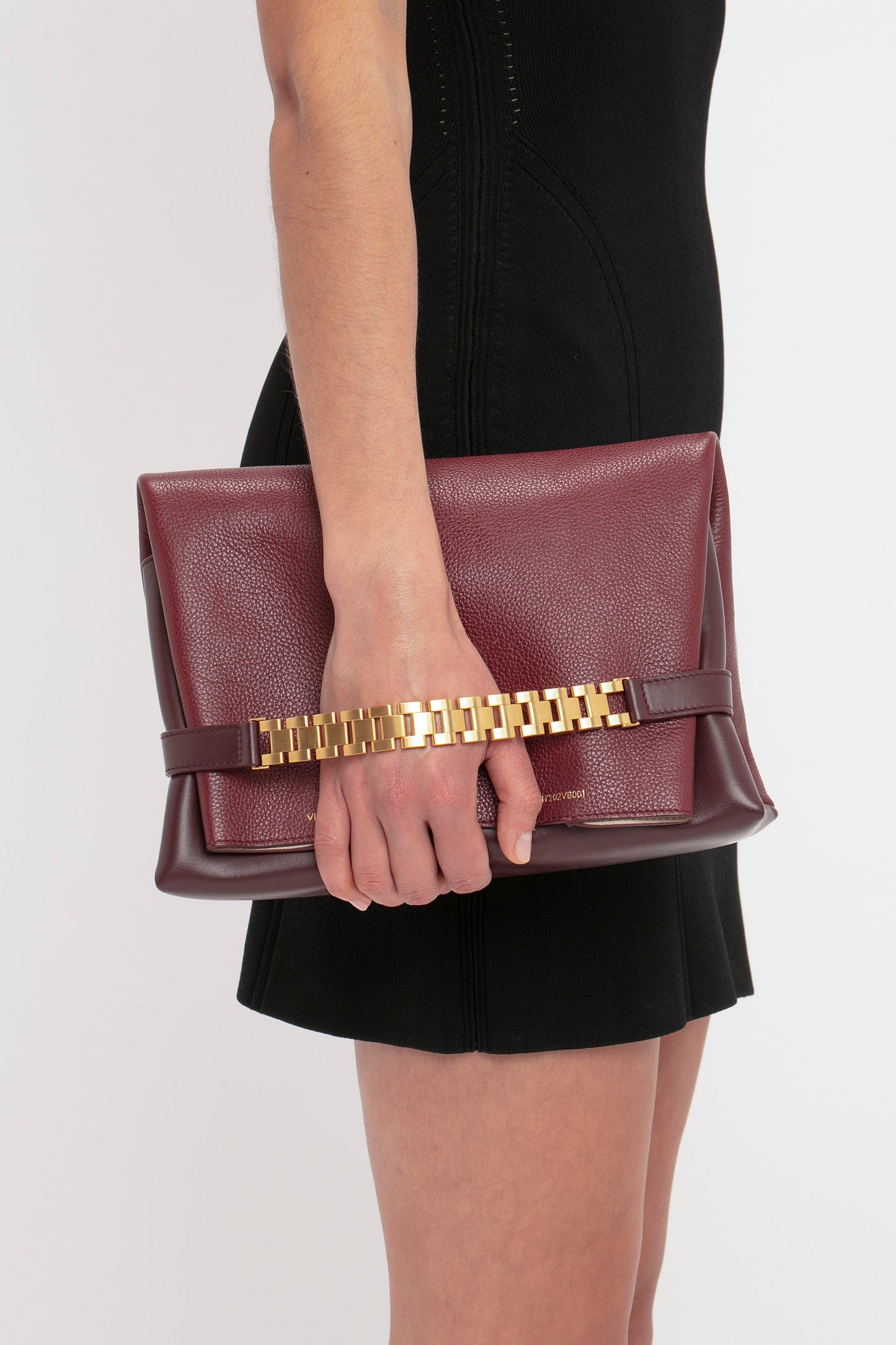 A person in the VB Body Compact Cap Sleeve Mini Dress in Black by Victoria Beckham holds a burgundy clutch with a rectangular gold accent.