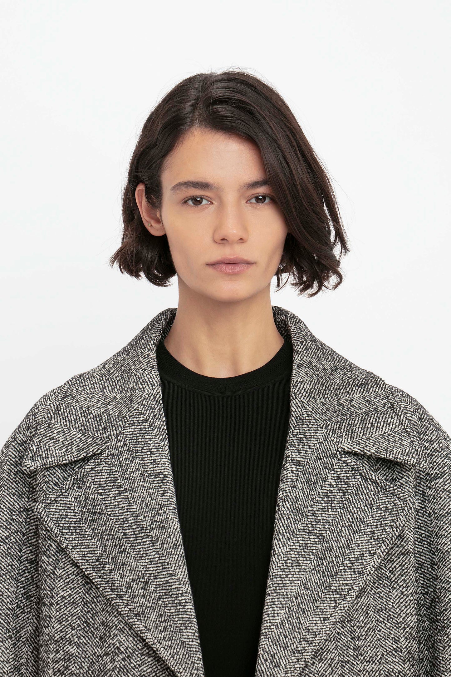 A person with short dark hair is wearing the VB Body Mini Dress in black from Victoria Beckham and a gray, wide-lapelled coat, looking directly at the camera against a plain background.