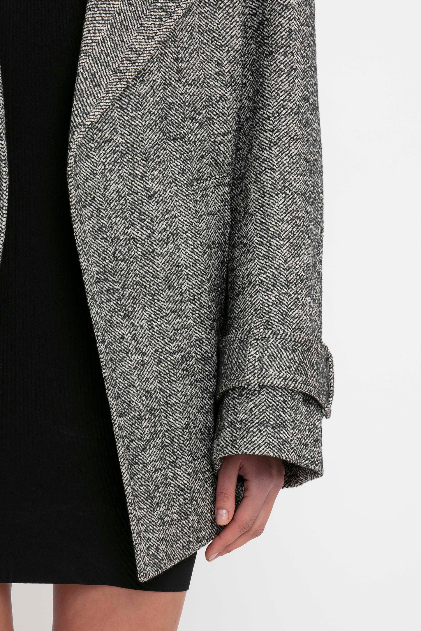 Close-up of a person wearing a grey herringbone coat over the VB Body Mini Dress in black by Victoria Beckham, with visible arm and hand at the coat’s hem.