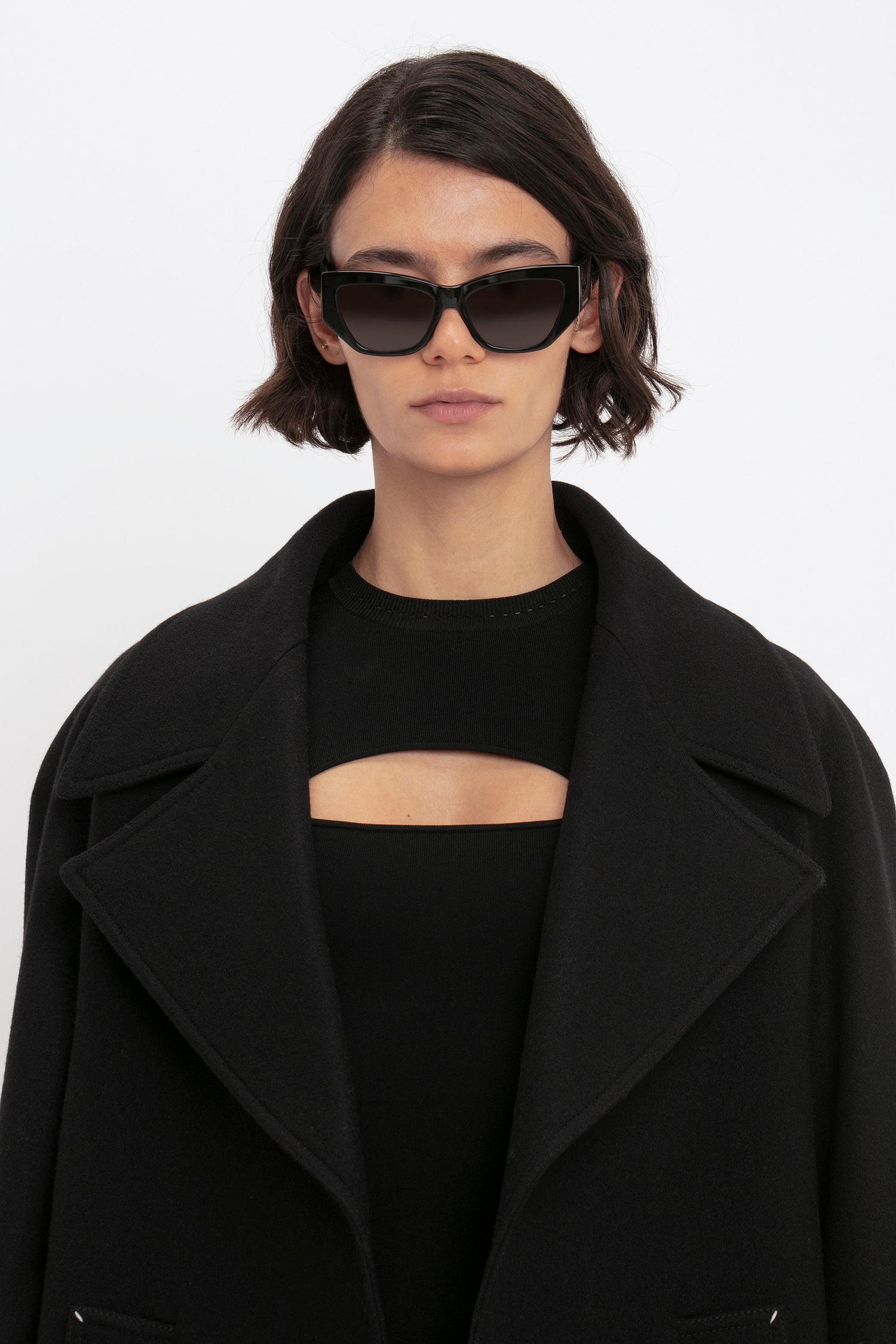 A person with short hair wears black sunglasses and the Victoria Beckham VB Body Cut Out Midi Dress in Black, posing against a plain white background.