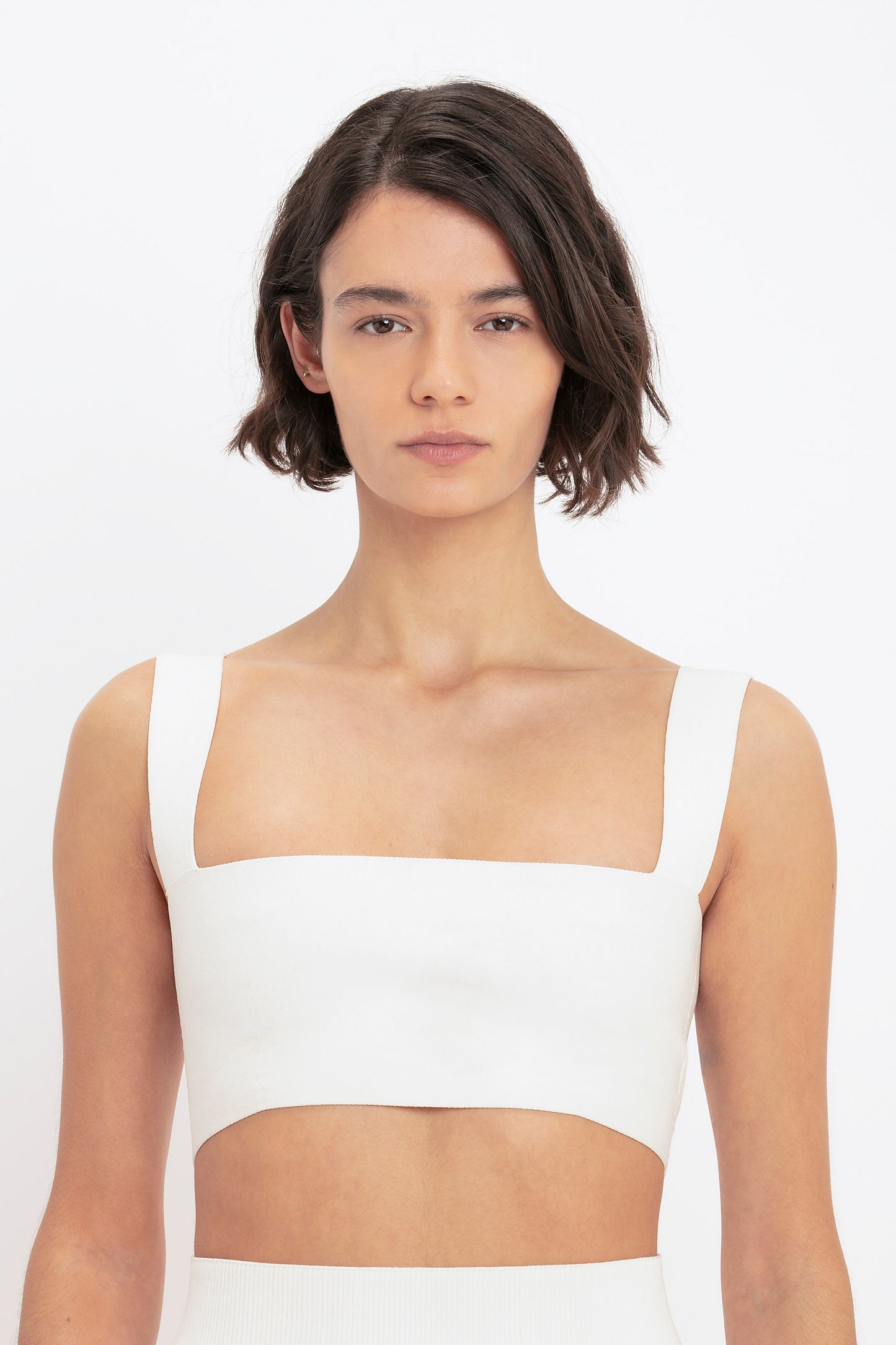 A woman with short dark hair wears the Victoria Beckham VB Body Strap Bandeau Top in White, standing against a plain background.