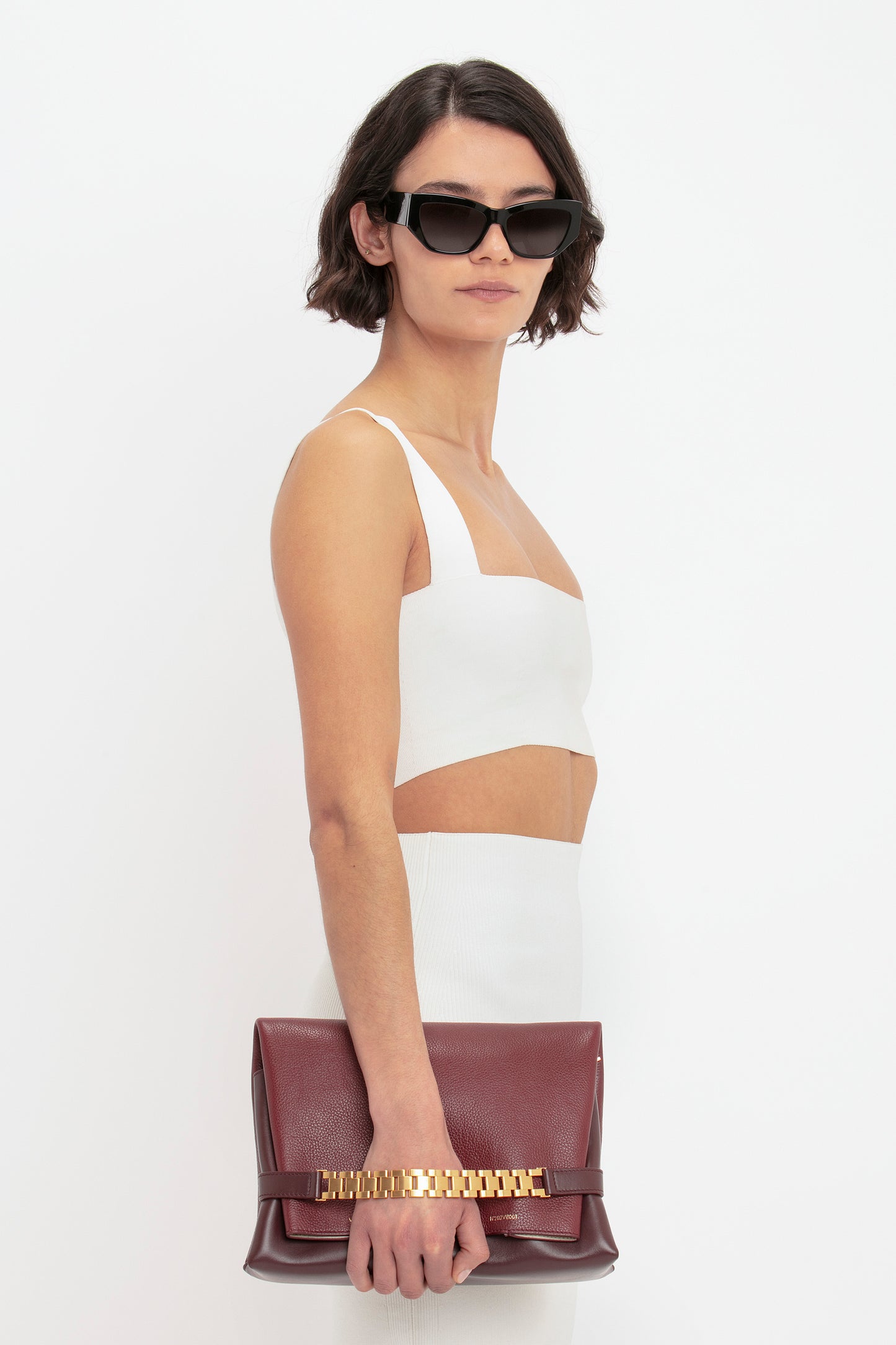 A person with short dark hair wearing sunglasses, a white VB Body Strap Bandeau Top by Victoria Beckham, and white pants, holding a maroon clutch bag with a gold chain detail.