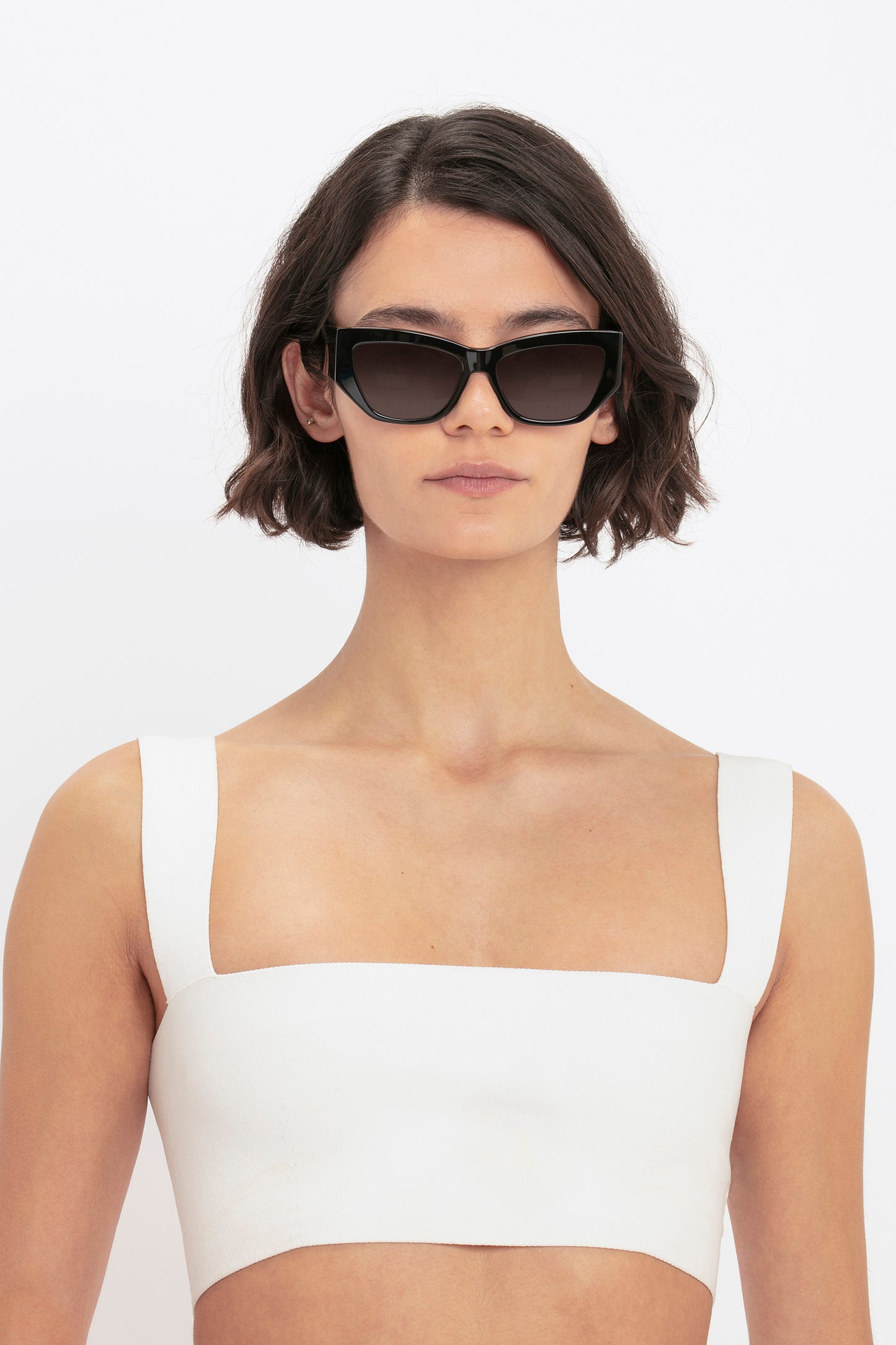 A woman with short, wavy hair wears black cat-eye sunglasses and the VB Body Strap Bandeau Top in White by Victoria Beckham against a plain white background.