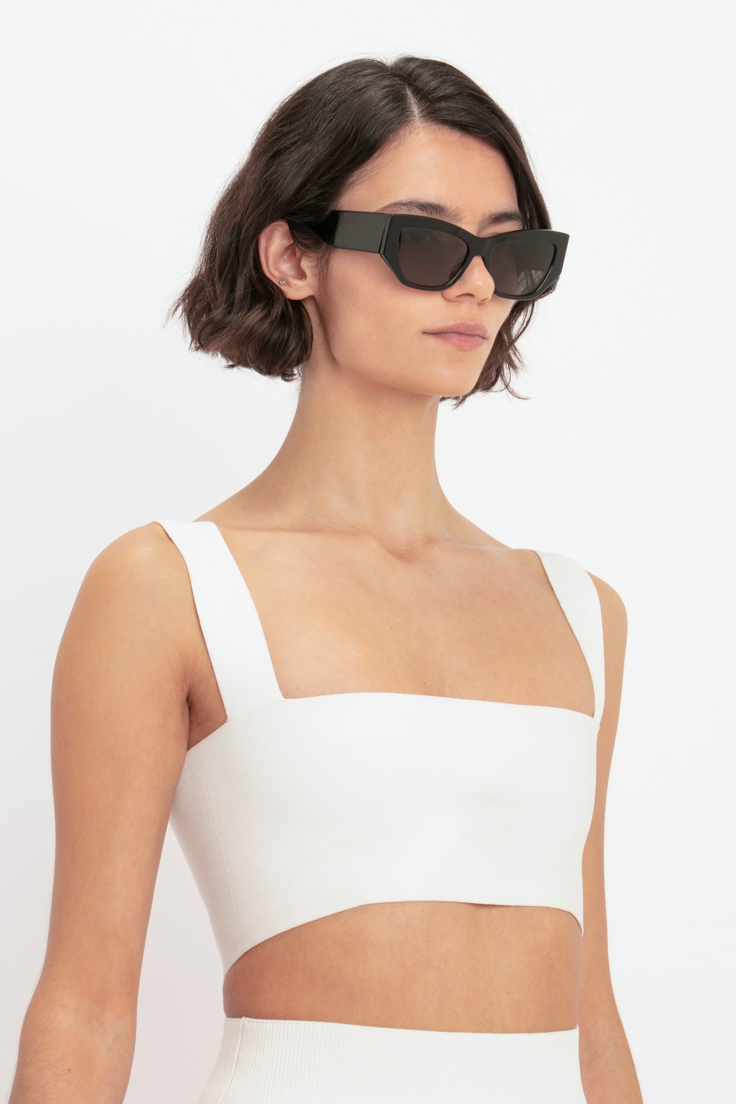 A person with short, dark hair wears the VB Body Strap Bandeau Top in White by Victoria Beckham and black rectangular sunglasses against a plain background.