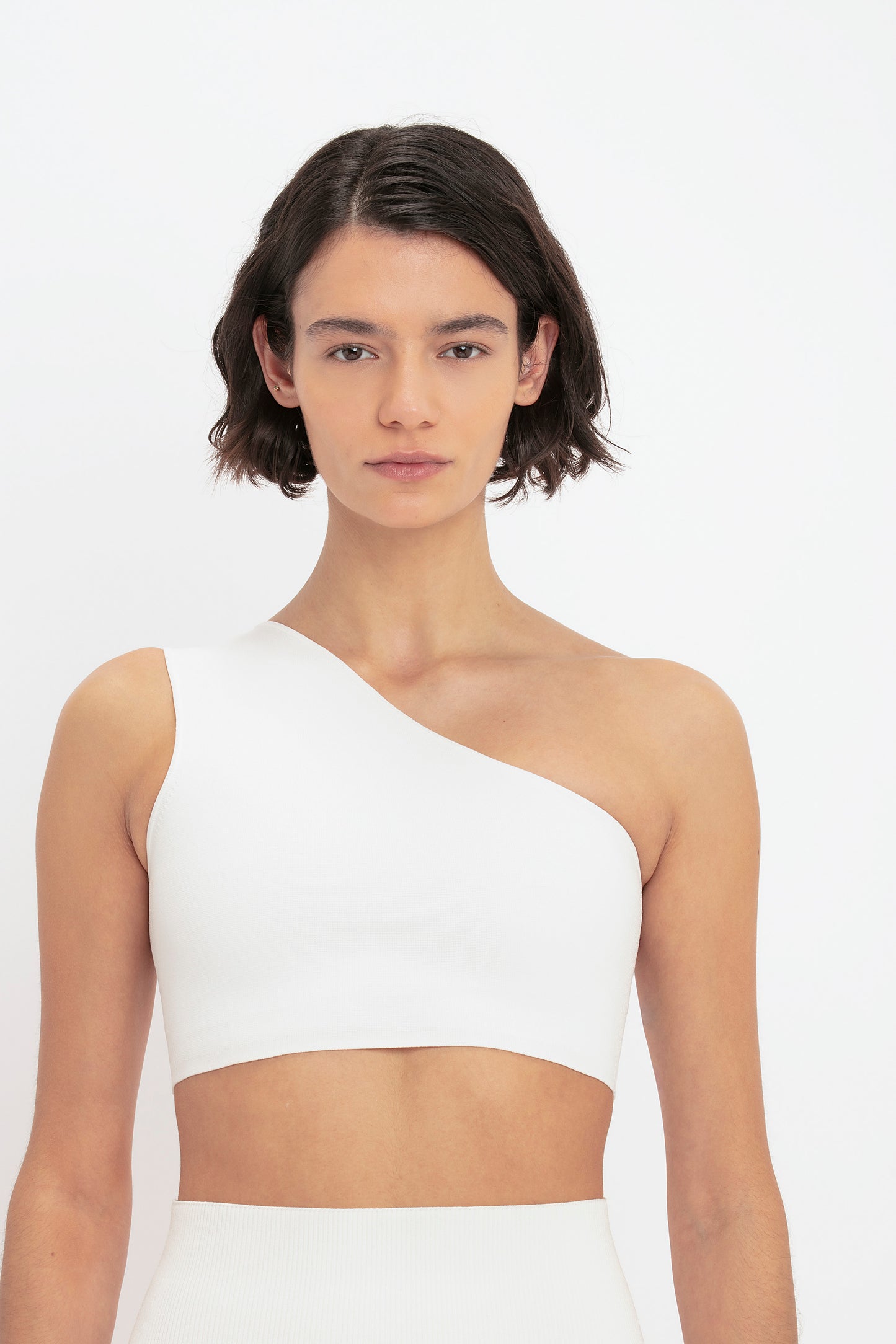 A person with short, dark hair wears the VB Body One Shoulder Crop Top in White by Victoria Beckham against a plain background.