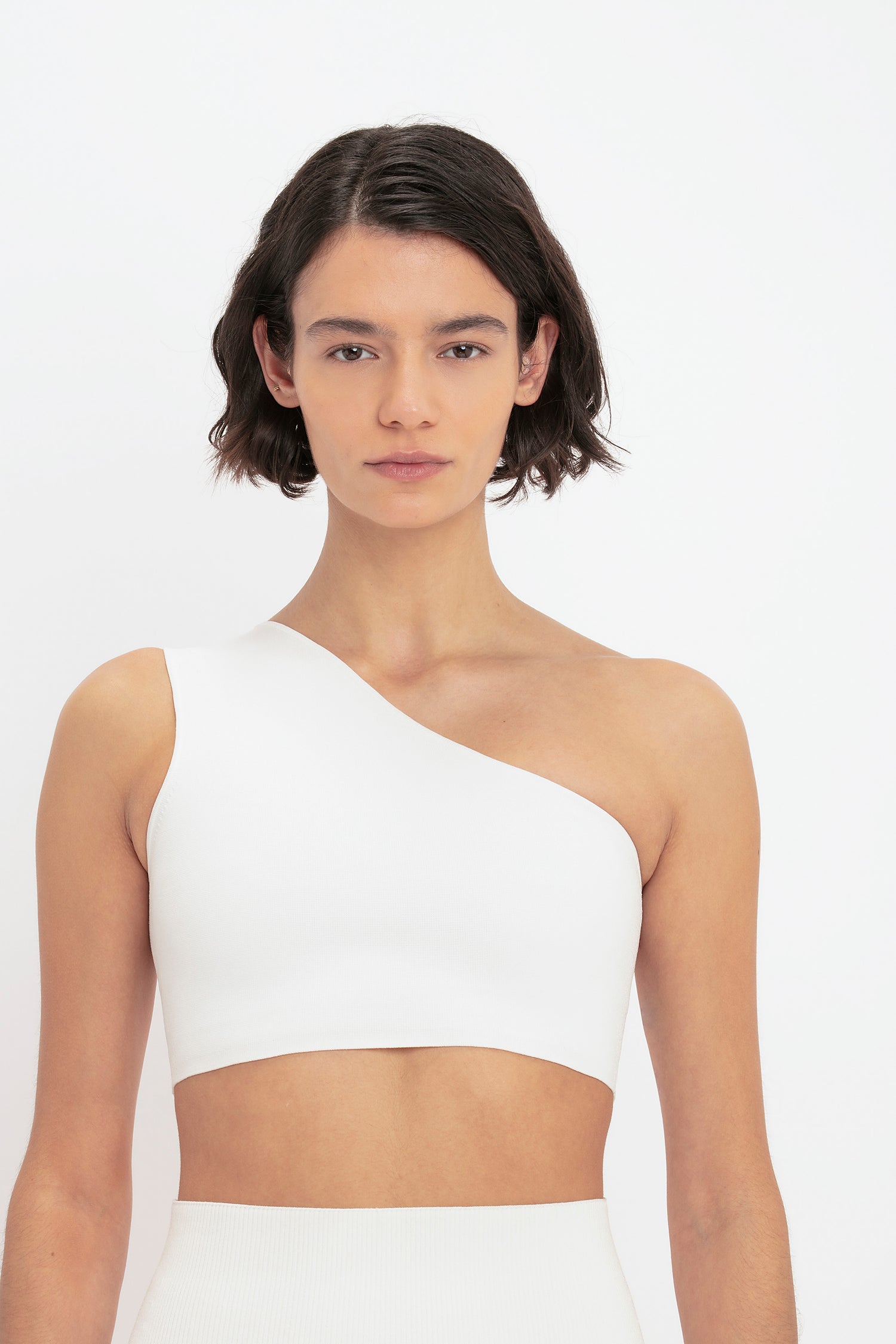 A person with short, dark hair wears the VB Body One Shoulder Crop Top in White by Victoria Beckham against a plain background.