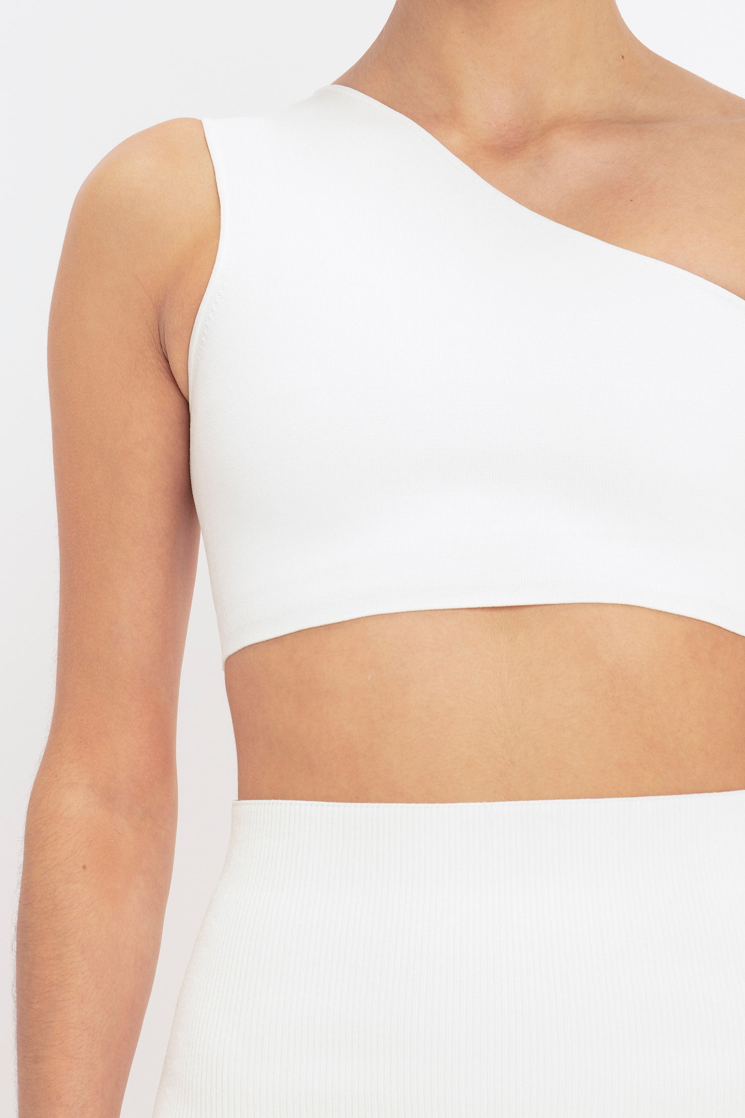 A person is shown from the neck to waist wearing a white one-shoulder crop top and the VB Body Fitted Midi Skirt in White by Victoria Beckham.