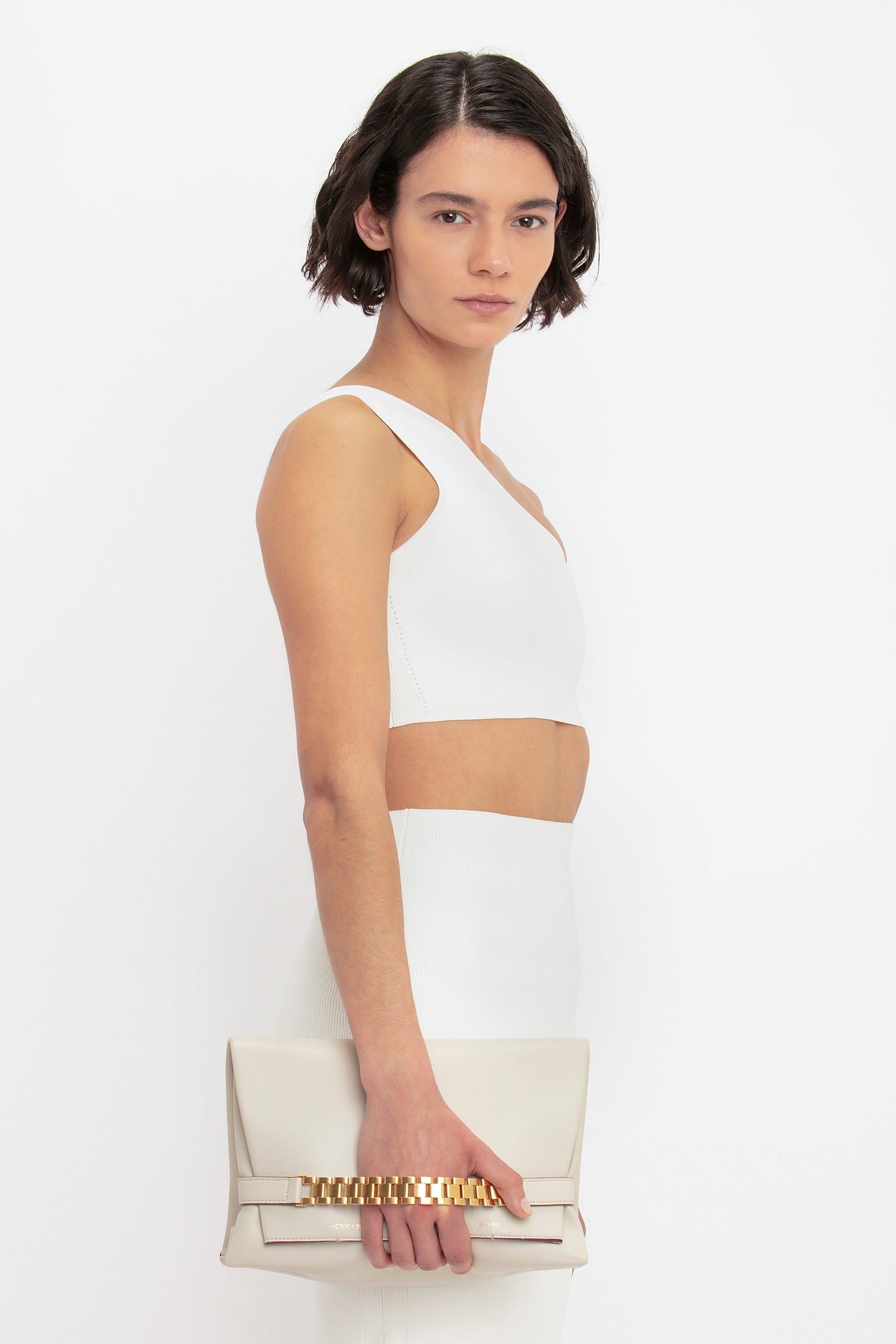 A person with short dark hair is wearing a white sleeveless top and a Victoria Beckham VB Body Fitted Midi Skirt in White, holding a beige clutch bag with a gold chain detail.