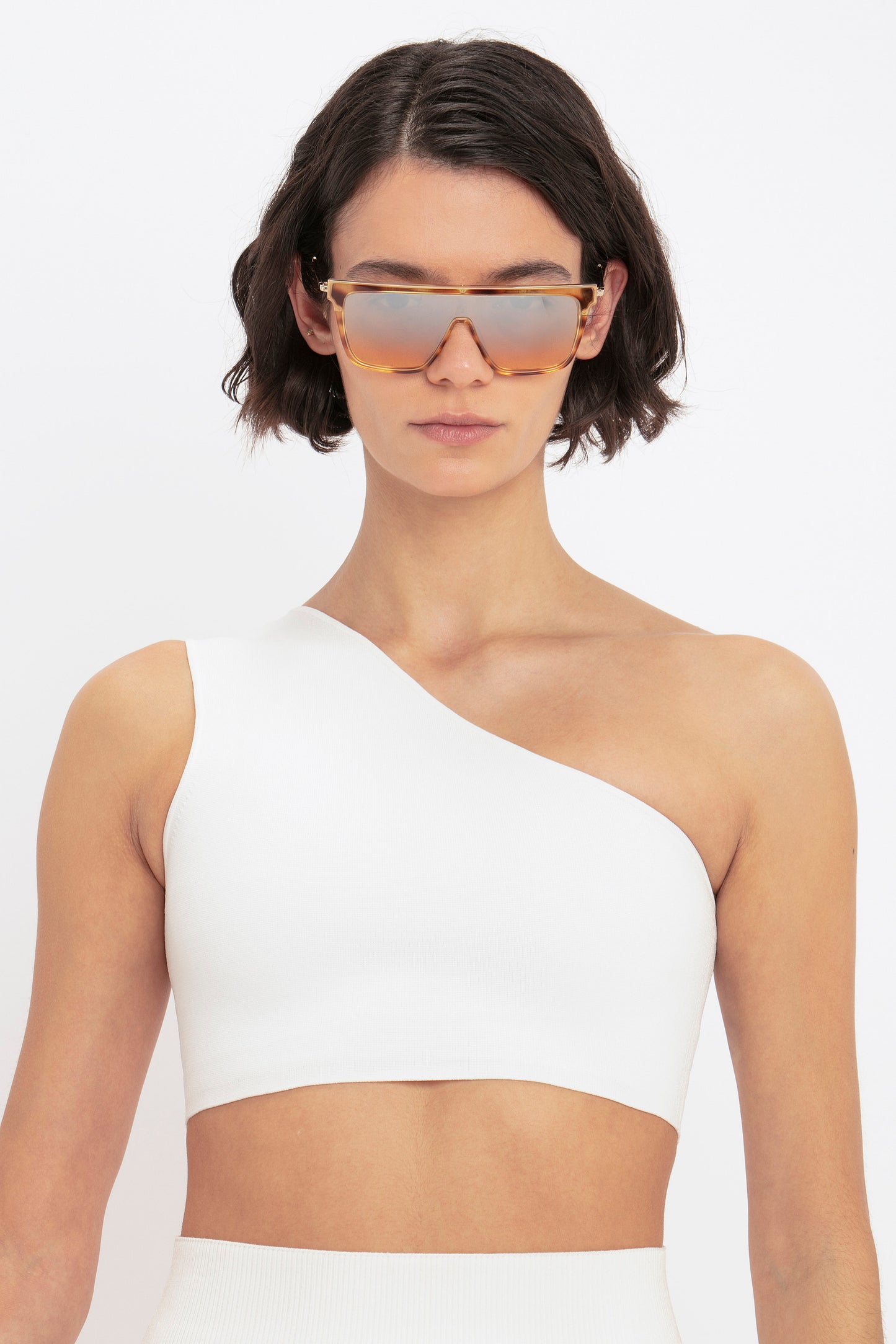 A person with short dark hair wears large, rectangular sunglasses and the VB Body One Shoulder Crop Top from Victoria Beckham against a plain white background.
