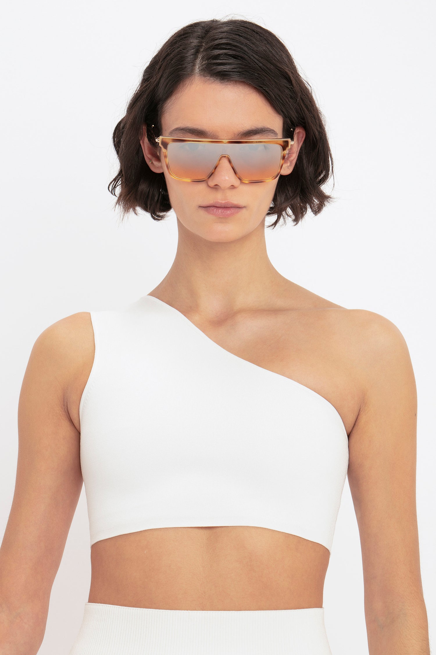 A person with short dark hair wears large, rectangular sunglasses and the VB Body One Shoulder Crop Top from Victoria Beckham against a plain white background.