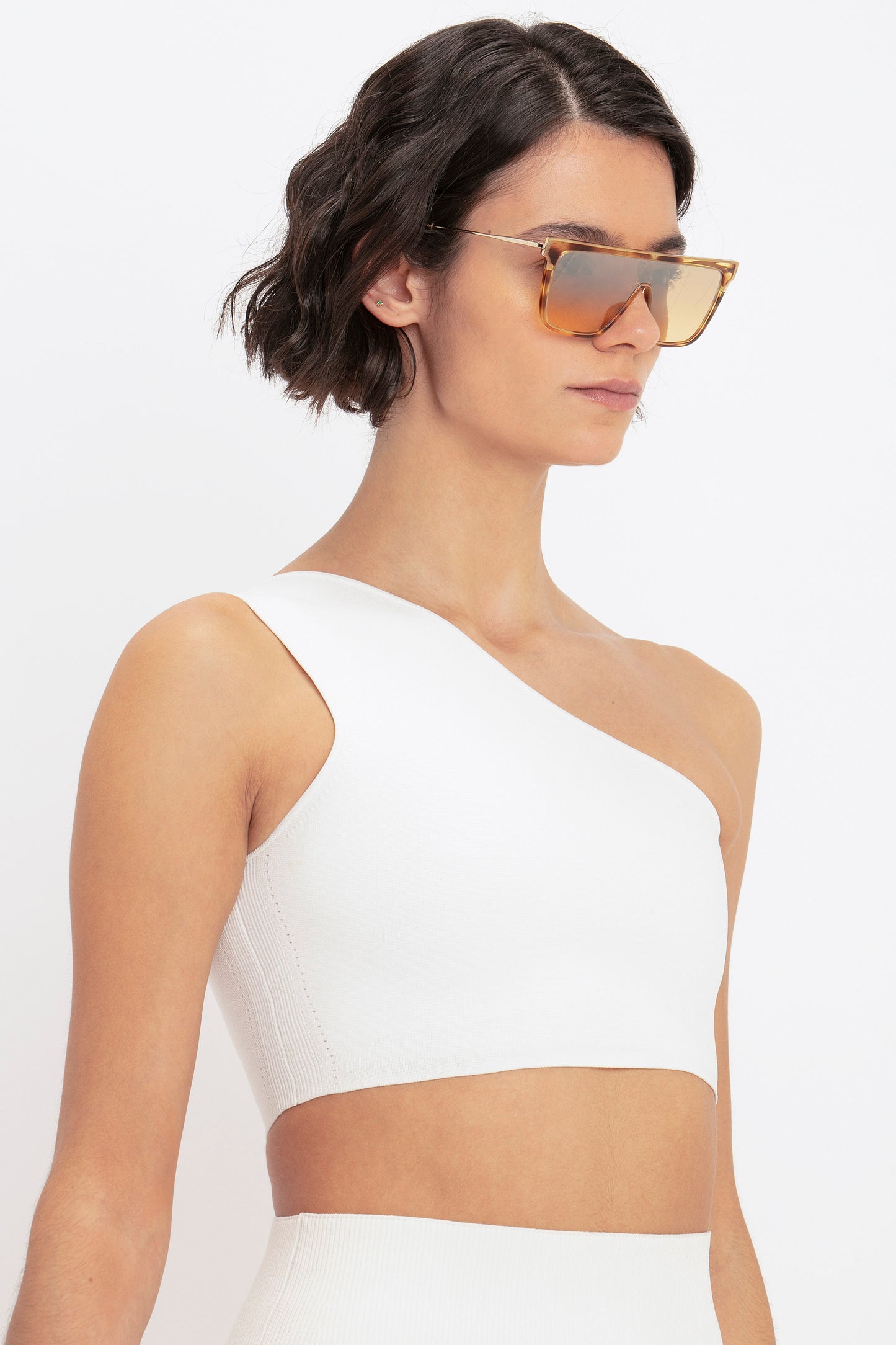 A person with short, dark hair wearing large sunglasses and a VB Body One Shoulder Crop Top in White by Victoria Beckham, looking to the side.