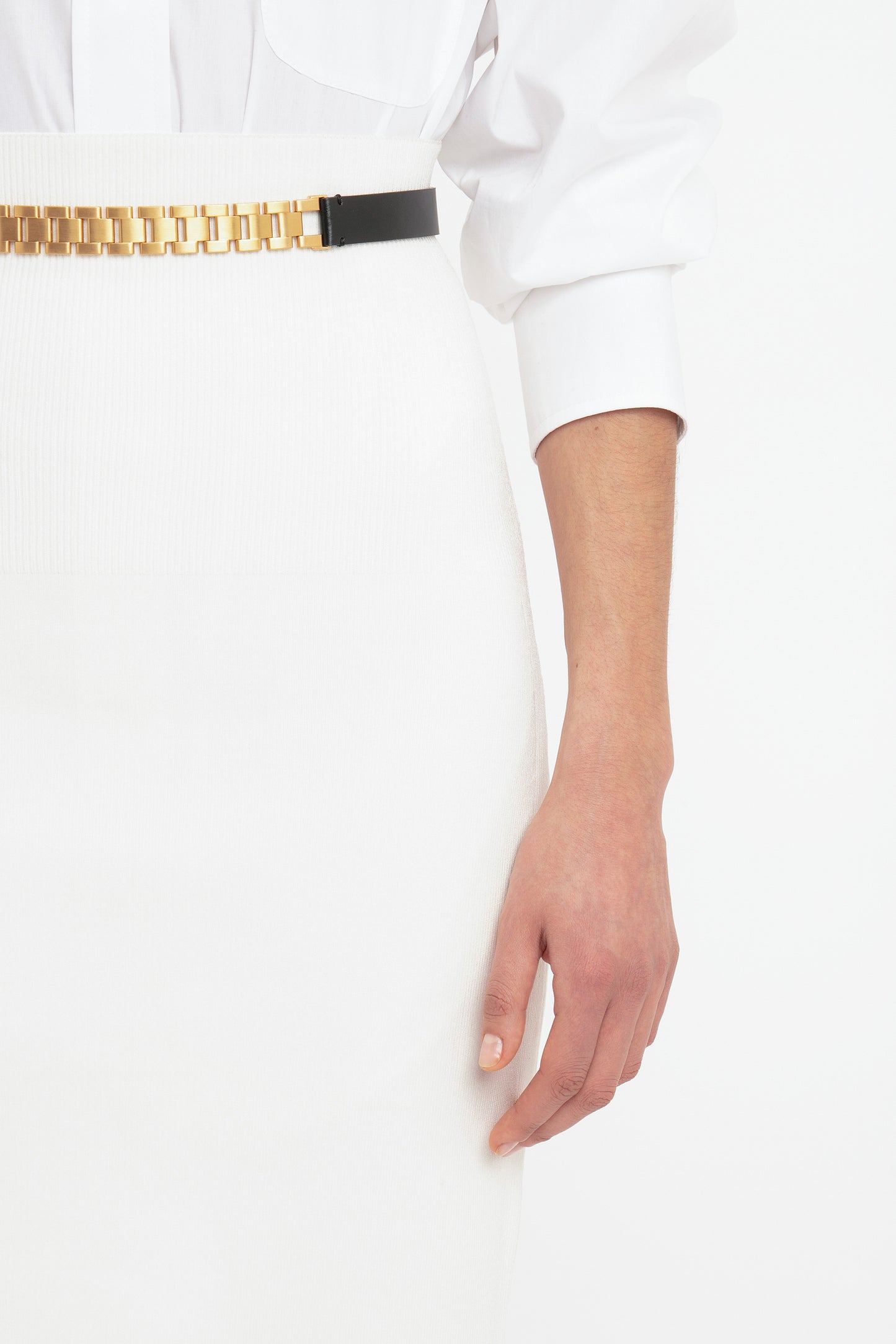 Close-up of a person wearing a white outfit featuring the VB Body Fitted Midi Skirt in White by Victoria Beckham, accented with a gold chain belt and showing their hand resting by their side.