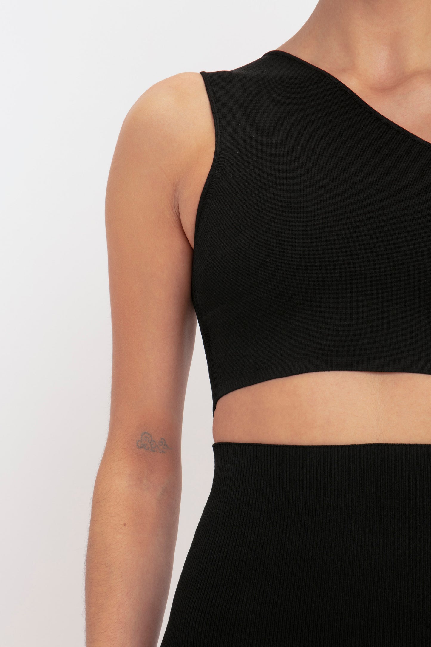 A person wearing the VB Body One Shoulder Crop Top in Black by Victoria Beckham, with a small tattoo of three circles visible on their upper arm.