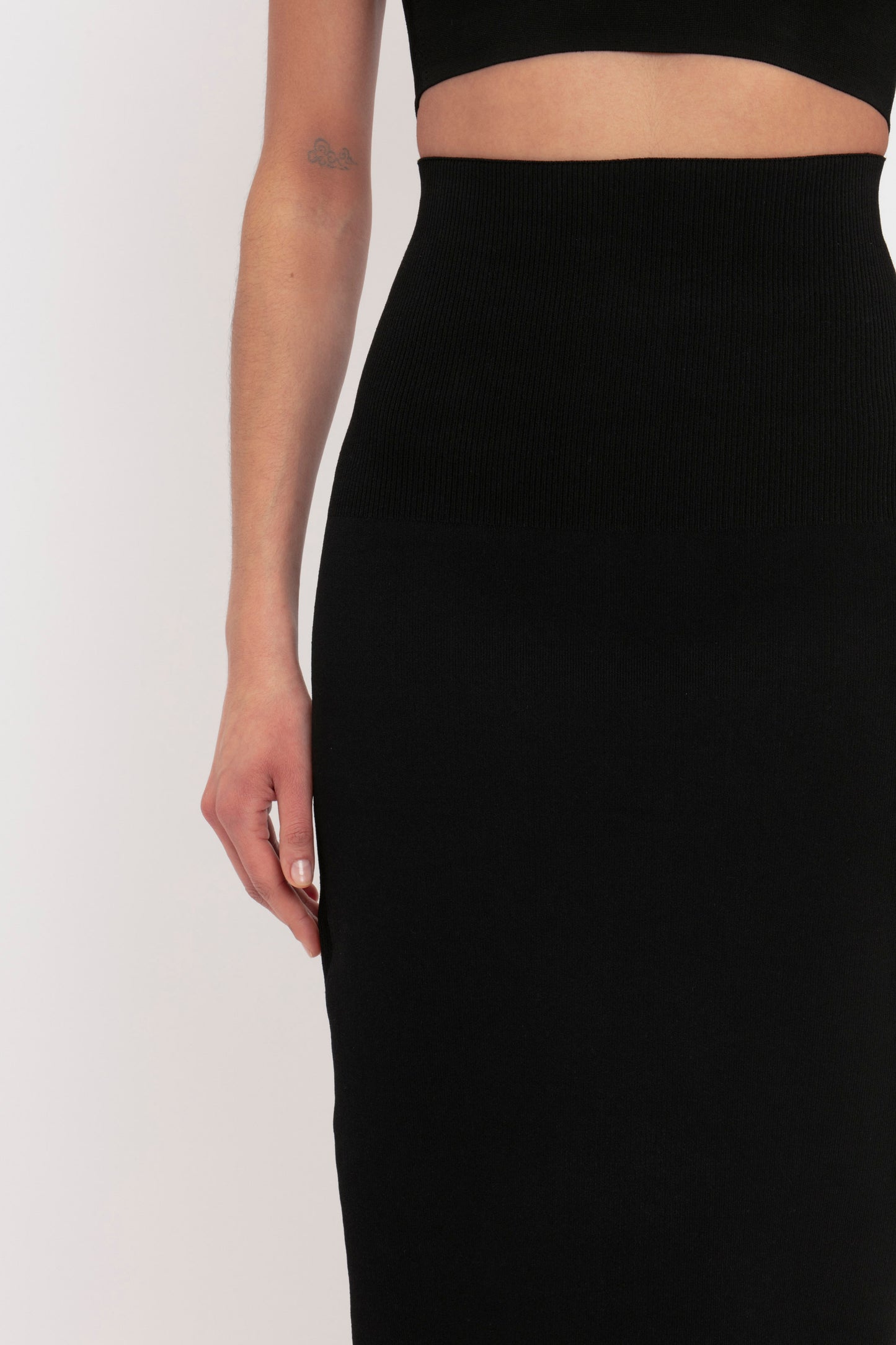 A person is wearing the VB Body Fitted Midi Skirt in Black by Victoria Beckham and a black crop top. The photo captures their midsection and left arm, revealing a small tattoo on their upper arm.