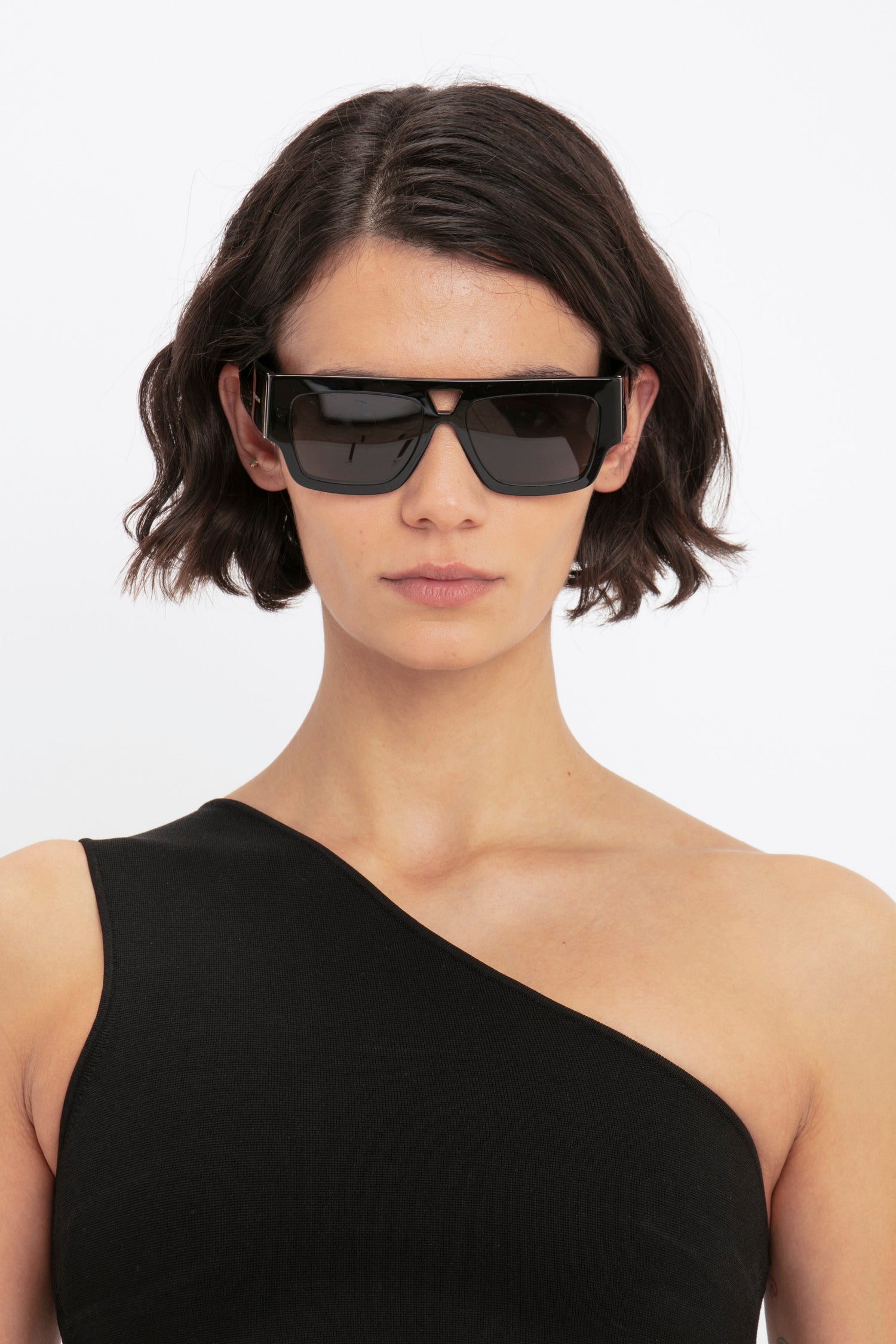 Person with short, dark hair wearing black sunglasses and the Victoria Beckham VB Body One Shoulder Crop Top in black, posing against a plain white background.