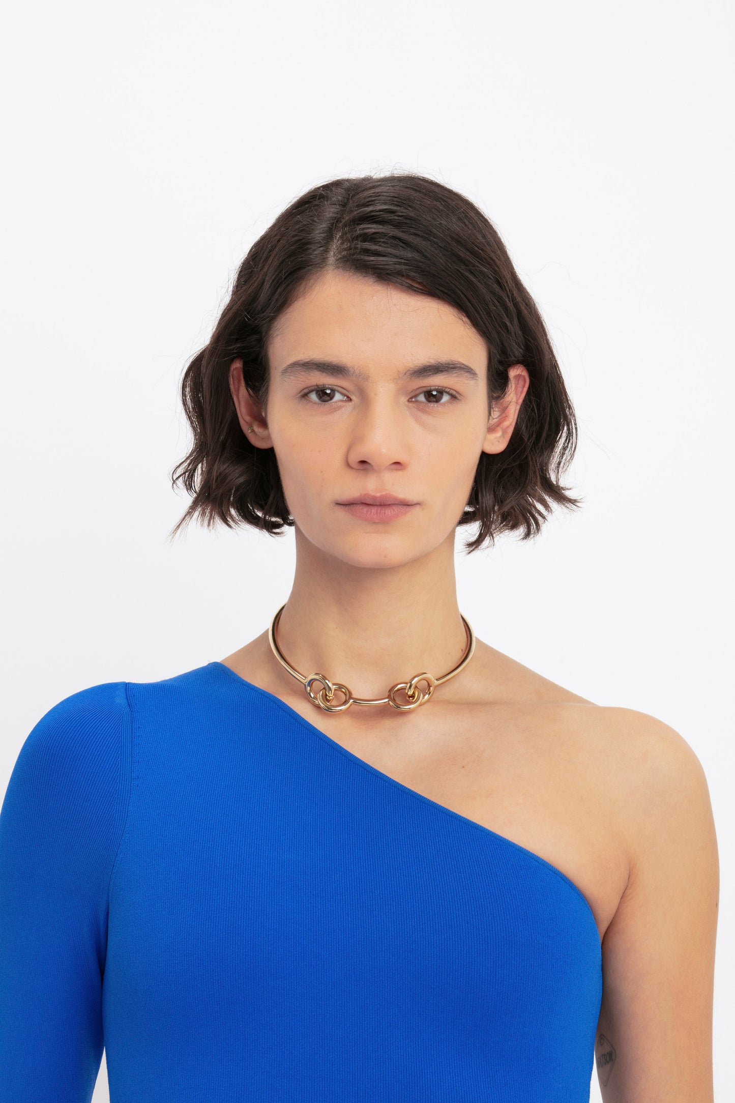 A person with short brown hair is wearing the VB Body One Shoulder Midi Dress in Cobalt by Victoria Beckham along with a gold choker necklace, posing against a plain white background.
