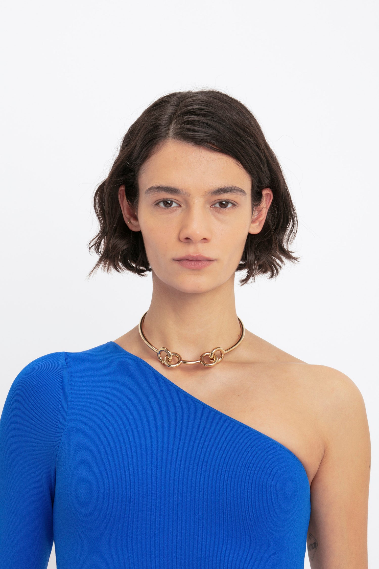 A person with short brown hair is wearing the VB Body One Shoulder Midi Dress in Cobalt by Victoria Beckham along with a gold choker necklace, posing against a plain white background.