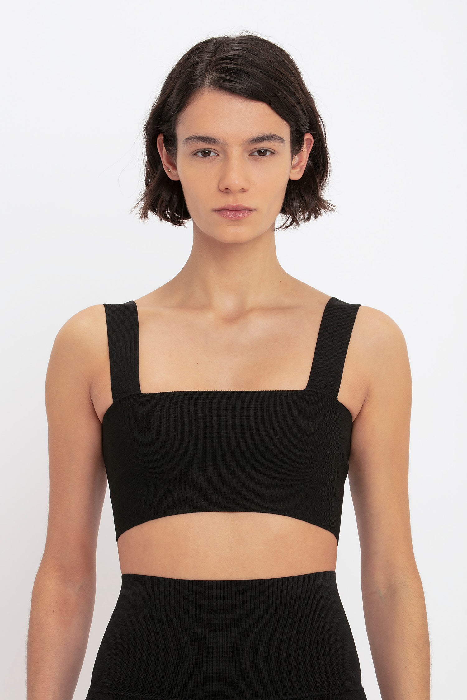 A person with short dark hair is wearing a black cropped tank top and looking directly at the camera. They are also sporting VB Body Split Front Leggings in Black from Victoria Beckham. The background is plain white.