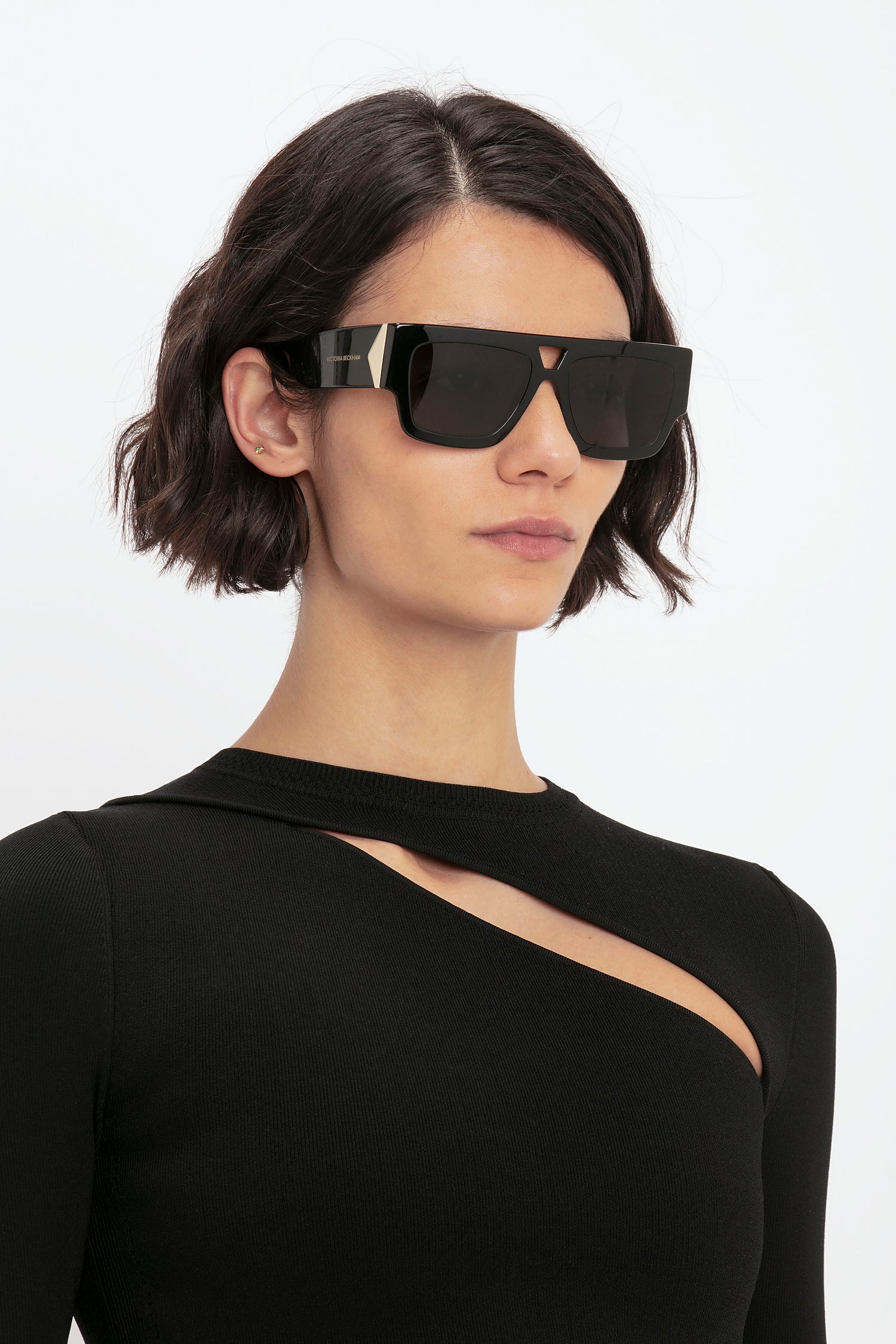 A woman with short dark hair wears large black sunglasses and the VB Body Front Slash Crew Neck Top in Black by Victoria Beckham.