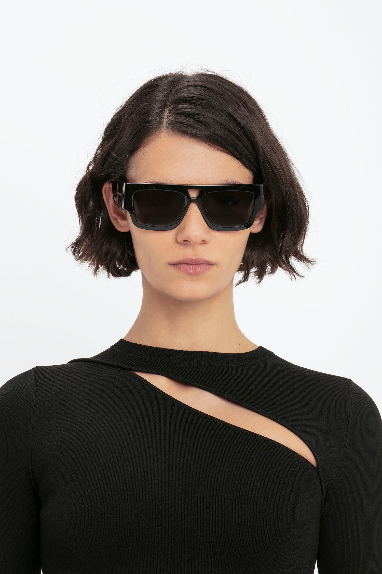 Individual with short, dark hair wearing black sunglasses and a black VB Body Front Slash Crew Neck Top by Victoria Beckham.