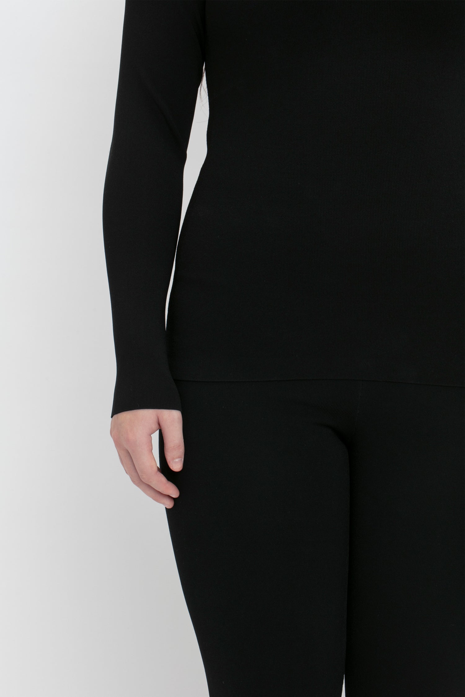 A person dressed in a black VB Body One Shoulder Top by Victoria Beckham and black pants, with their right hand partially visible at their side. The background is plain white.