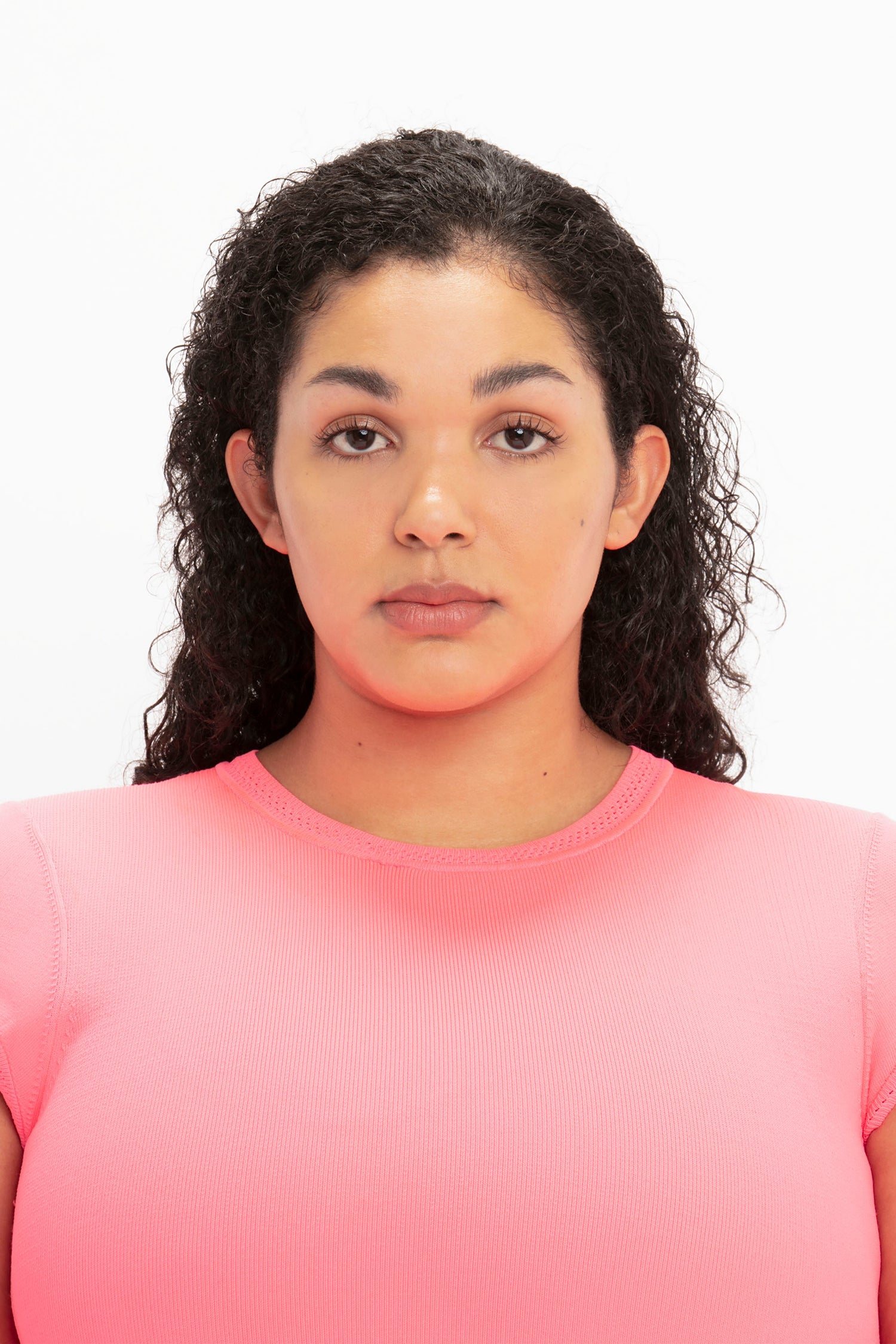 A person with curly hair wearing the Victoria Beckham VB Body Compact Cap Sleeve Mini Dress in Pink looks straight ahead with a neutral expression against a plain white background.