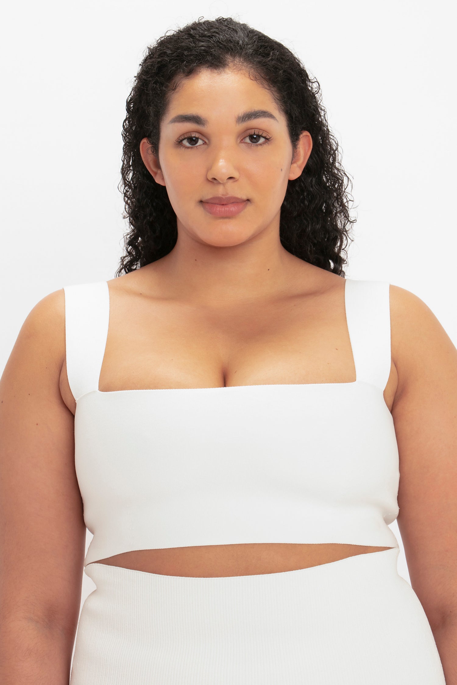 A woman with curly hair wears the Victoria Beckham VB Body Strap Bandeau Top in white, featuring wide straps and a cut-out detail under the bust, as she poses against a plain white background.