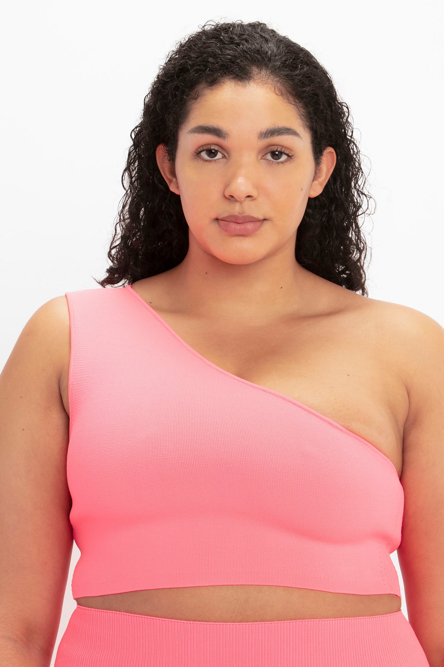 A person with curly hair wearing the Victoria Beckham VB Body One Shoulder Crop Top in pink is facing the camera with a neutral expression.
