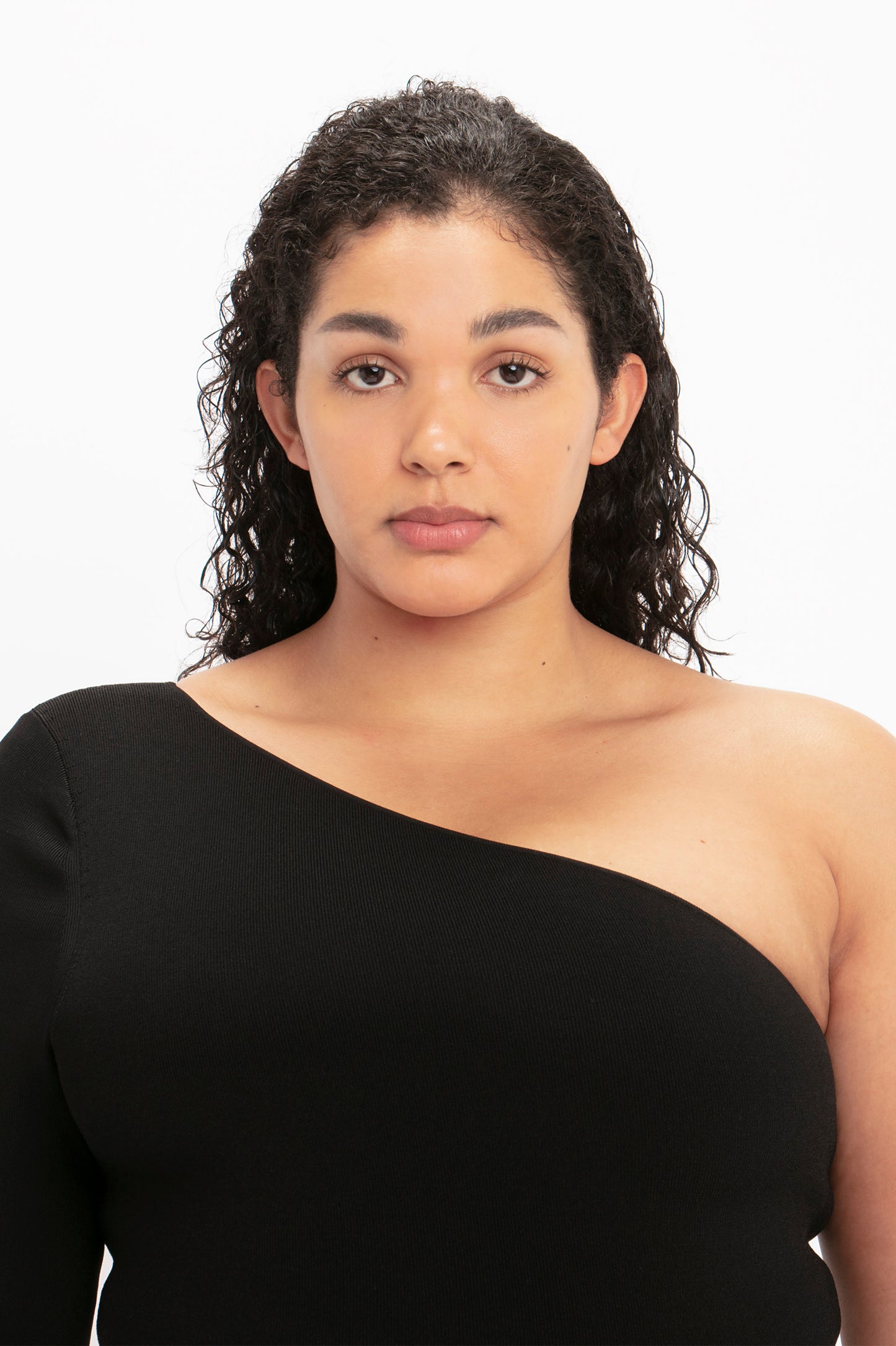 A person with curly dark hair, wearing the VB Body One Shoulder Midi Dress in Black by Victoria Beckham, looks directly at the camera against a plain white background.