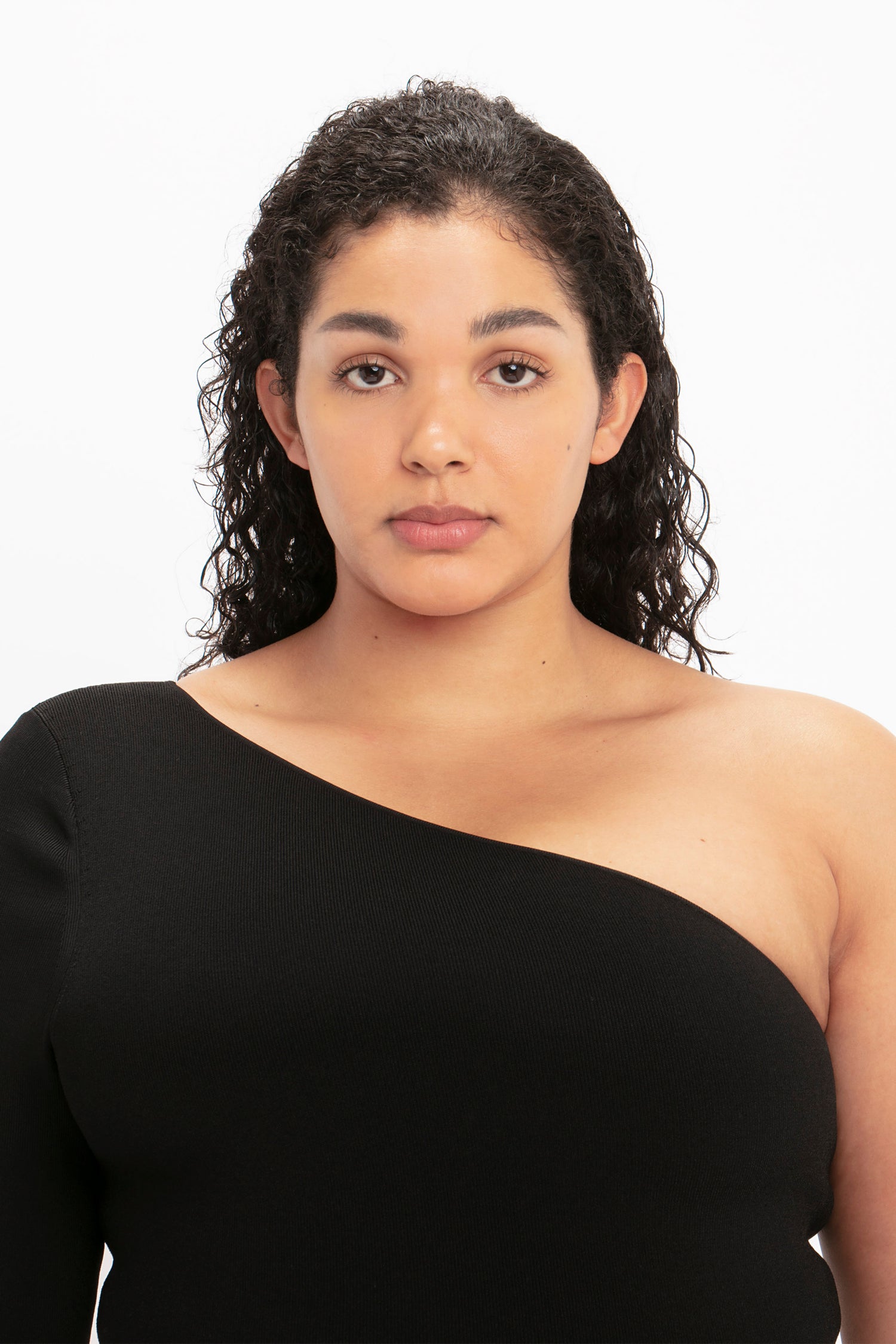 A person with curly dark hair, wearing the VB Body One Shoulder Midi Dress in Black by Victoria Beckham, looks directly at the camera against a plain white background.