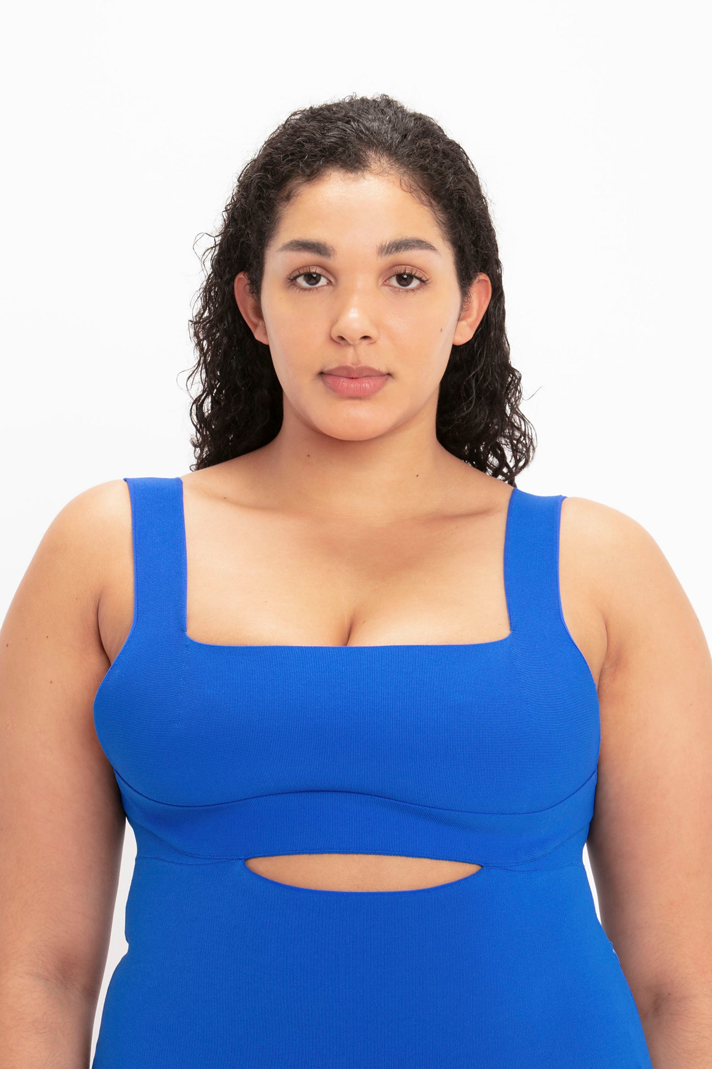 A person with curly hair is seen facing the camera against a plain white background, donning the VB Body Cut Out Midi Dress in Cobalt by Victoria Beckham, which features a bright blue sleeveless design with a square neckline and cut-out detail beneath the bust.