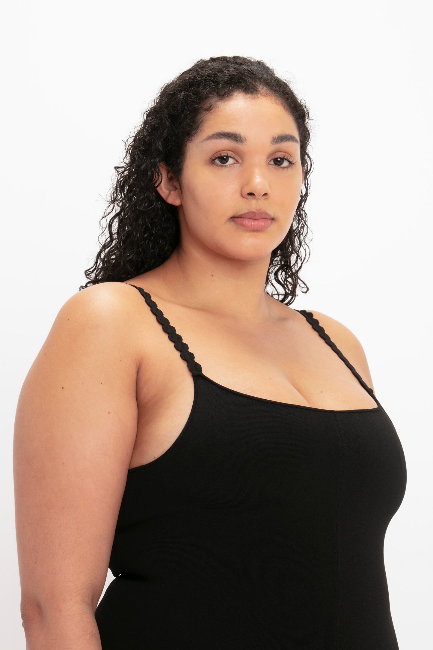 A woman with curly hair, wearing the VB Body Catsuit in Black by Victoria Beckham, is looking directly at the camera against a plain white background.