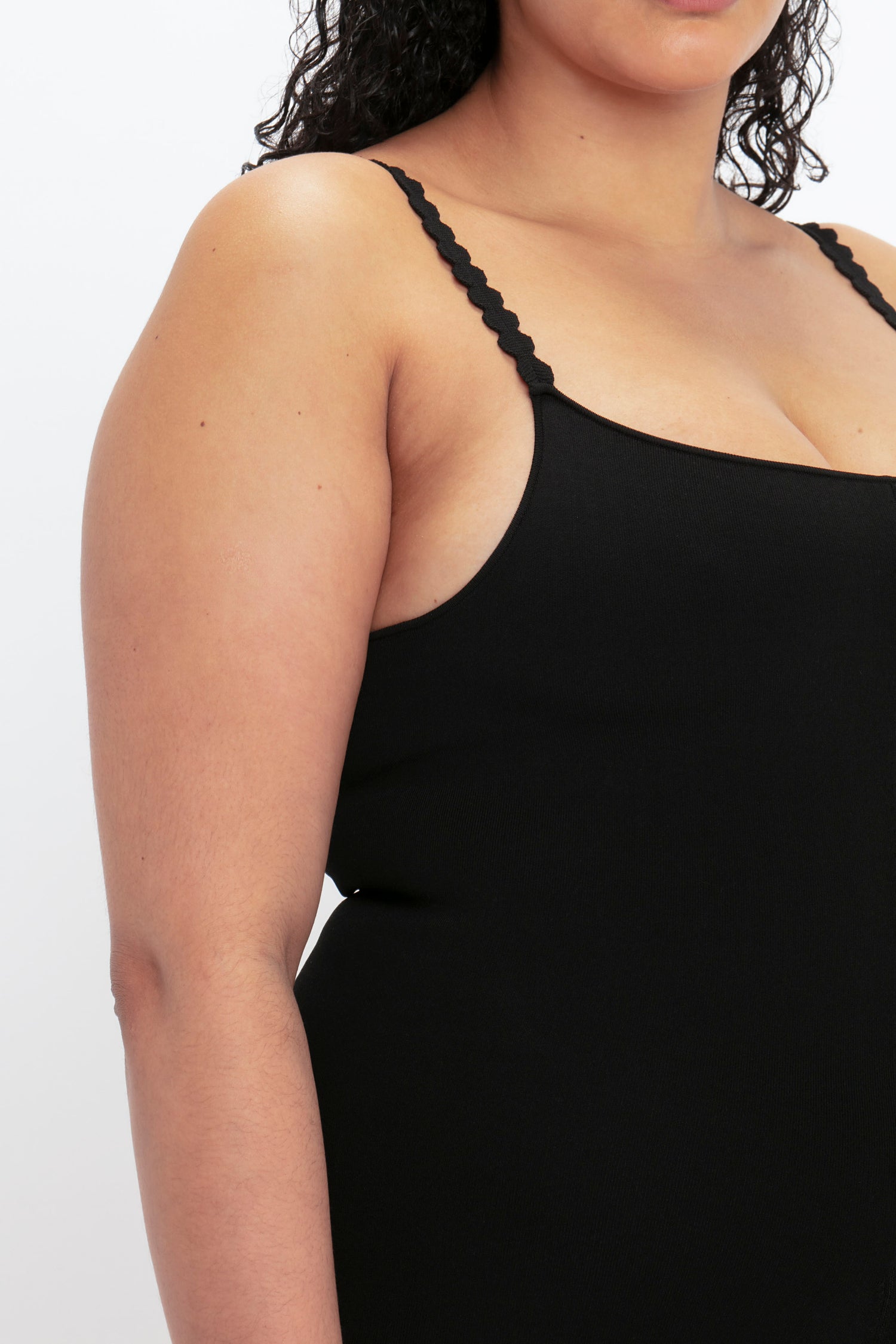 A person wearing the VB Body Catsuit in Black by Victoria Beckham with thin, scalloped straps. Only the left side of their upper body is visible against a plain background.