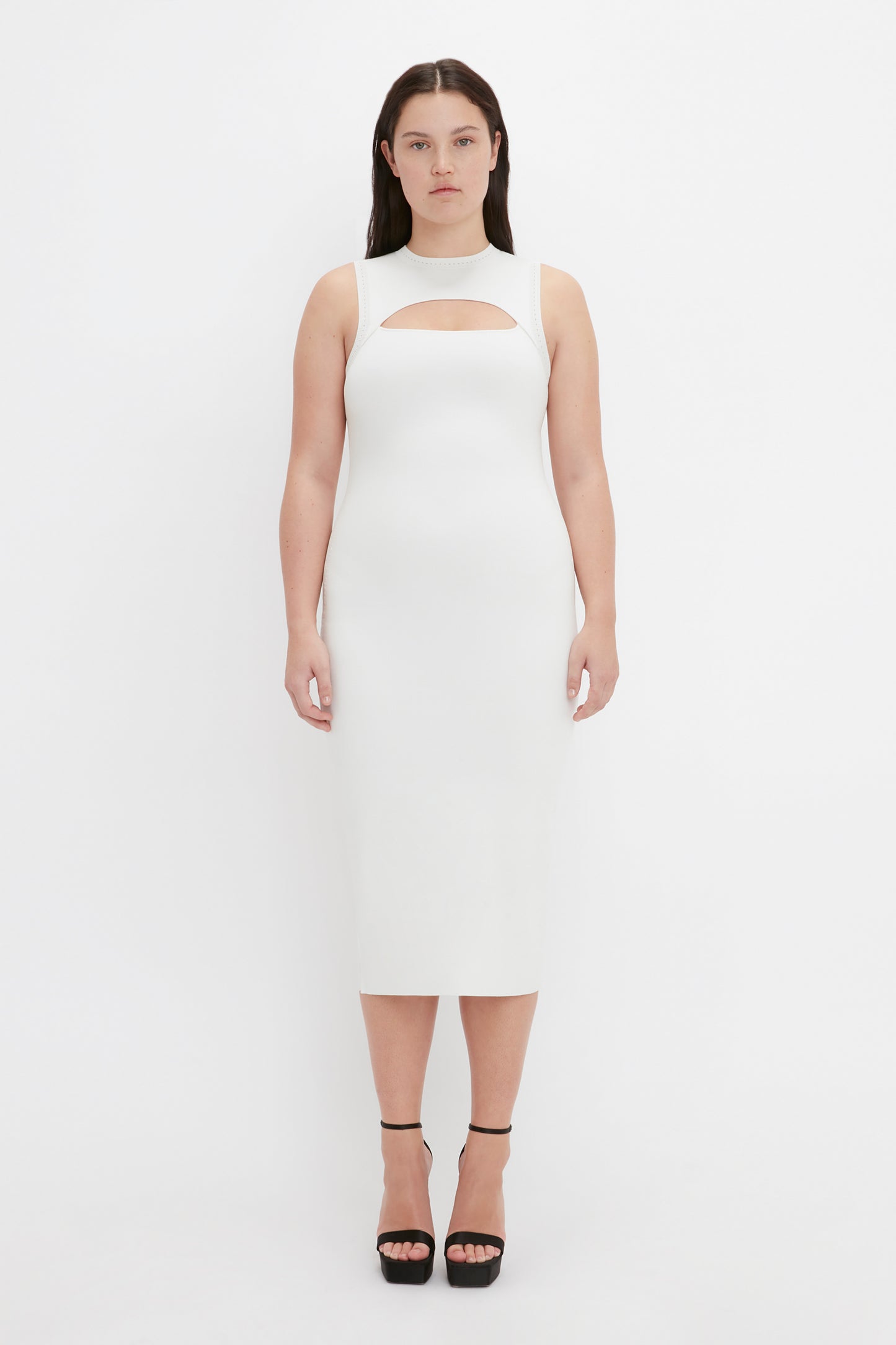 A woman with dark hair stands against a plain white background, wearing the VB Body Slash Front Midi Dress in White by Victoria Beckham, paired with black platform heels.