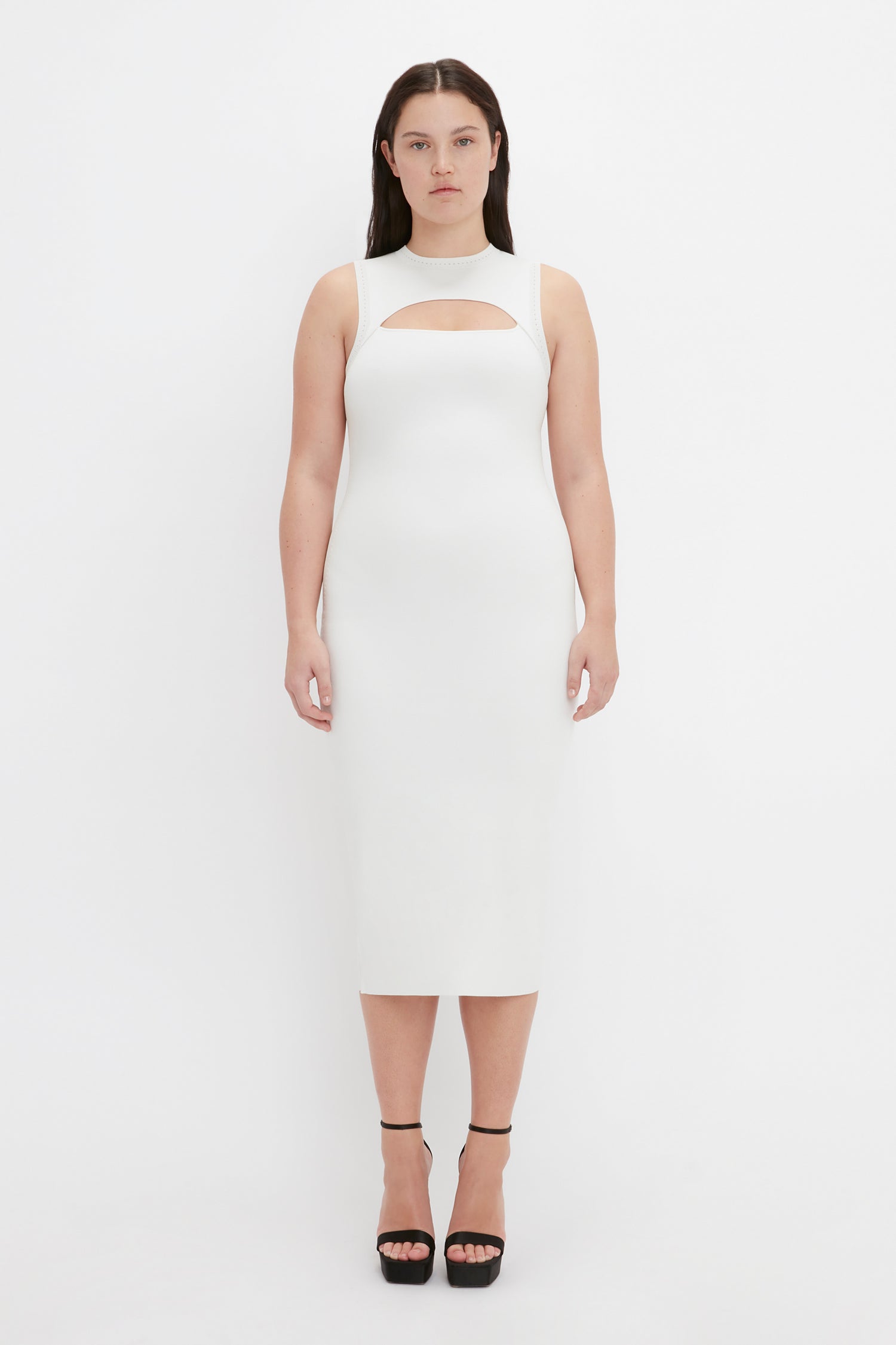 A woman with dark hair stands against a plain white background, wearing the VB Body Slash Front Midi Dress in White by Victoria Beckham, paired with black platform heels.