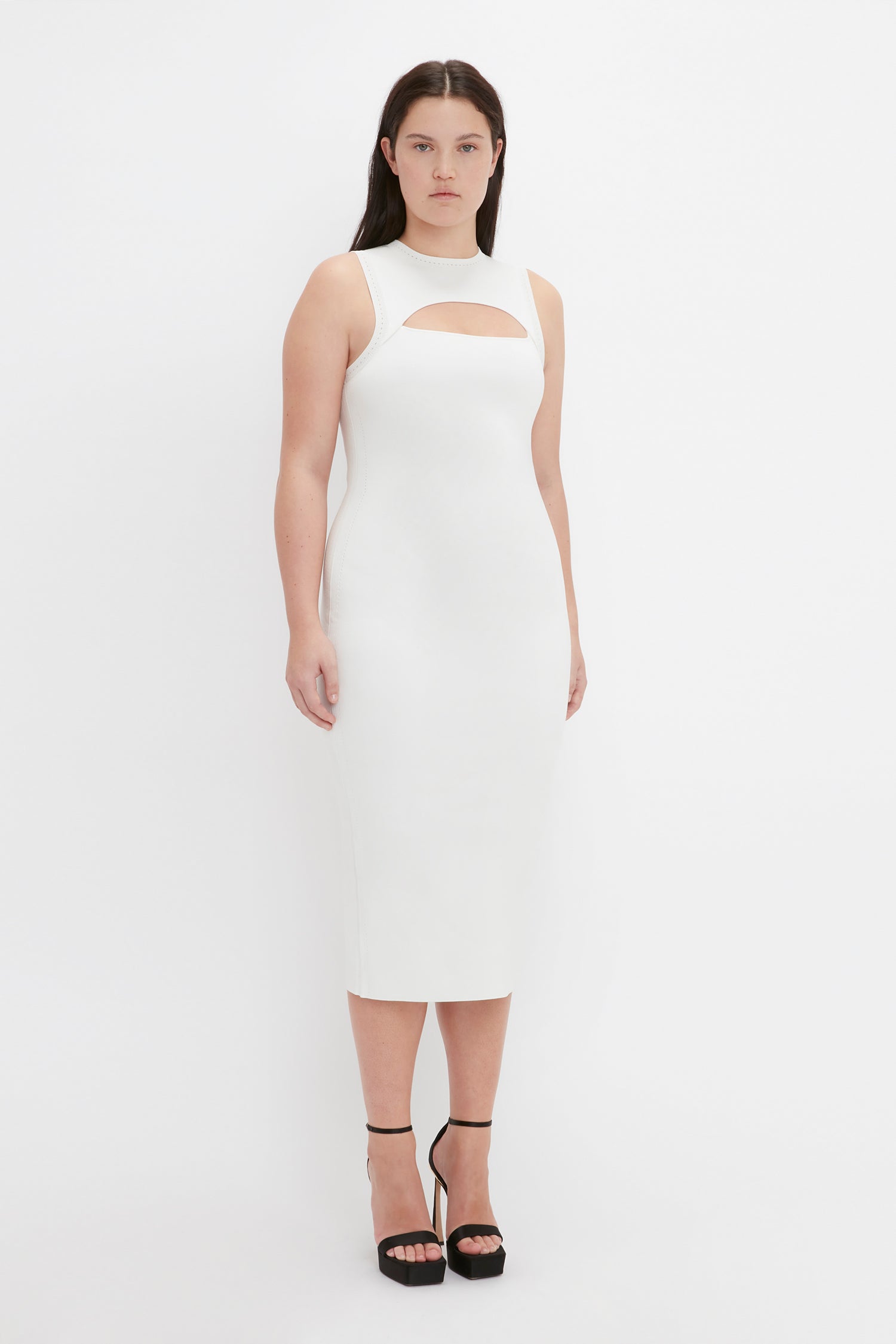 A woman with long dark hair is wearing the Victoria Beckham VB Body Slash Front Midi Dress in White and black high-heeled sandals, standing against a plain white background.
