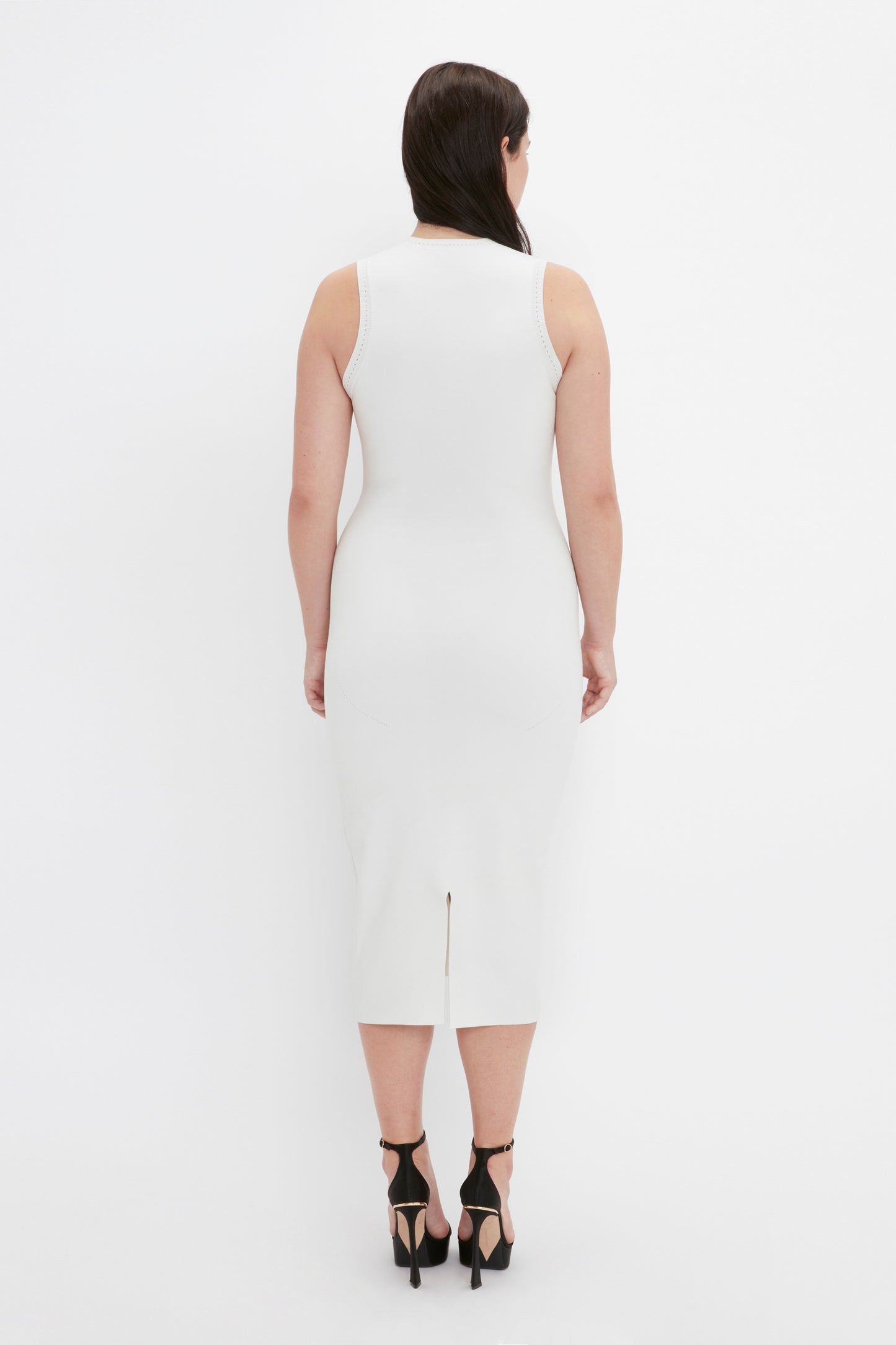 A woman with dark hair is facing away, wearing the VB Body Slash Front Midi Dress in White from Victoria Beckham, paired with black high-heeled shoes.