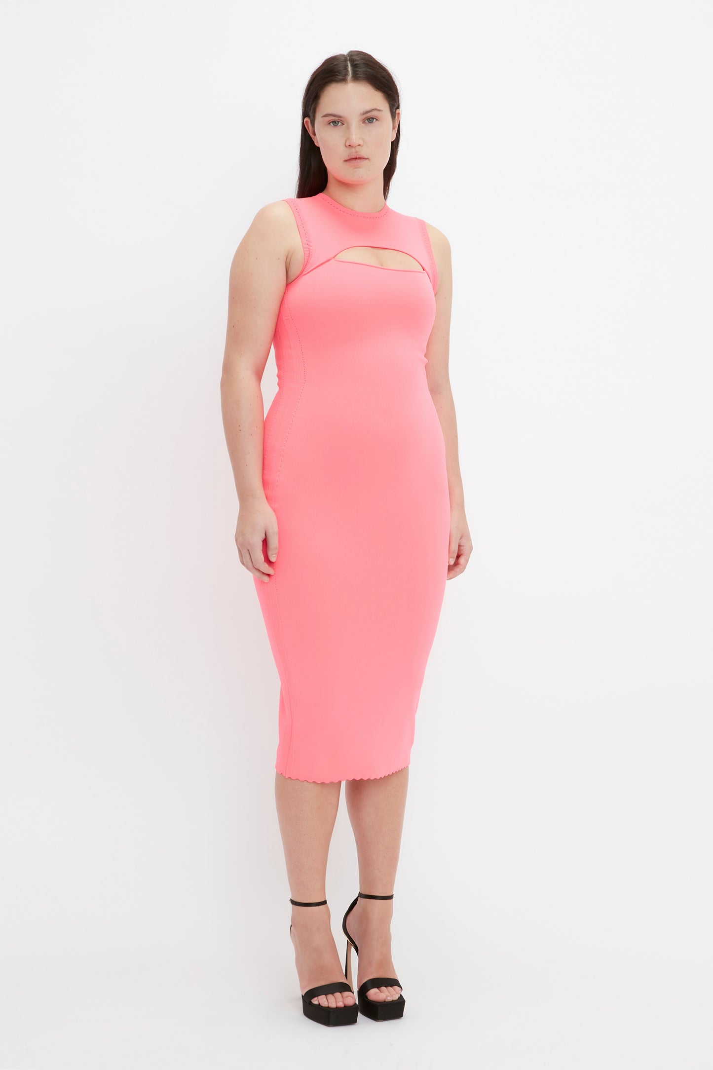 A person stands against a plain white background, wearing the Victoria Beckham VB Body Slash Front Midi Dress in Pink and black high-heeled shoes.