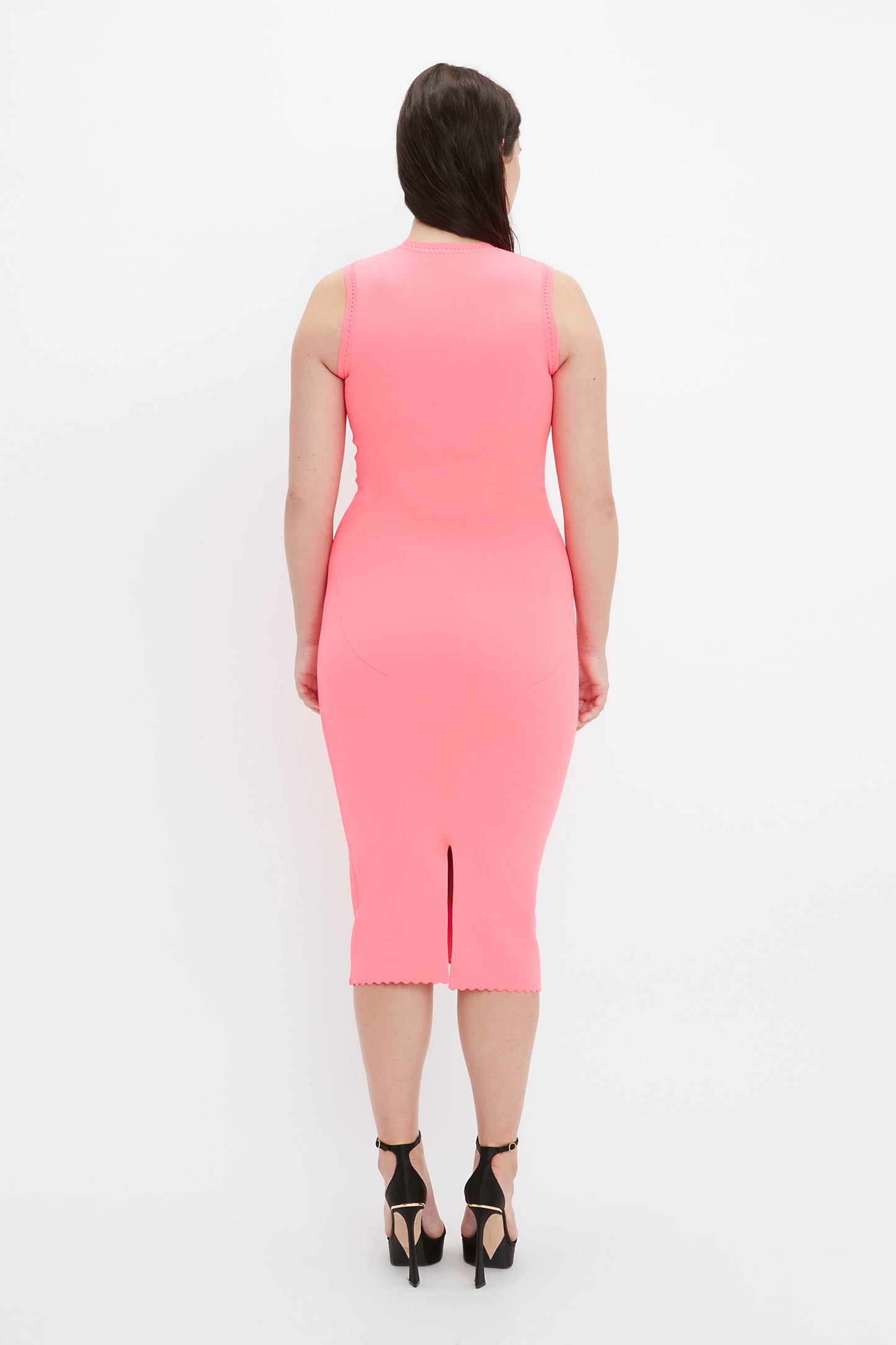 A woman in a Victoria Beckham VB Body Slash Front Midi Dress in pink and black high heels is facing away from the camera against a white background.