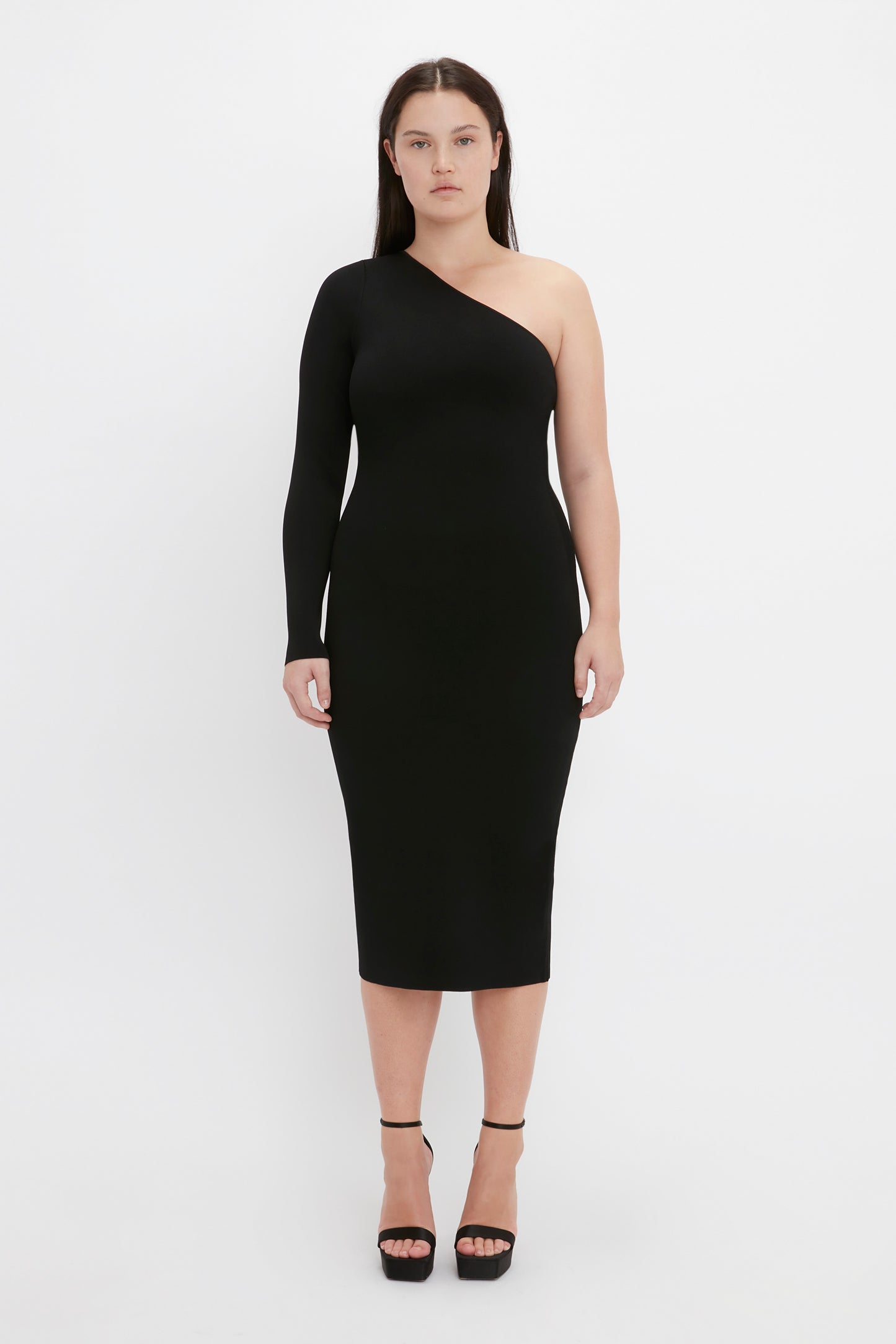 A person is wearing the VB Body One Shoulder Midi Dress in Black from Victoria Beckham, which features a fitted design with a single long sleeve. They are also sporting black high-heeled shoes against a plain white background.