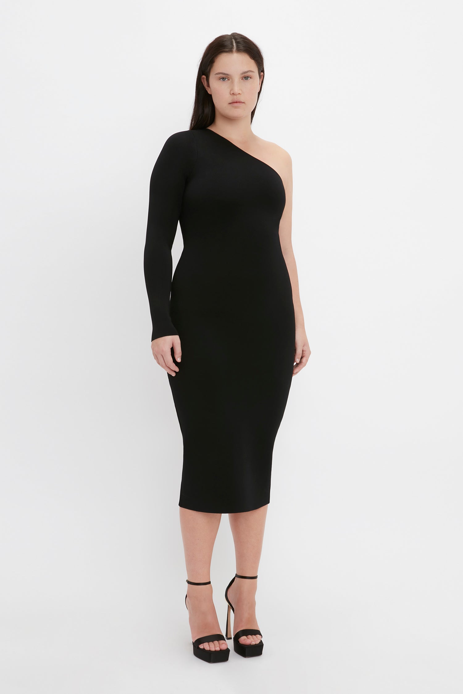 A woman stands against a plain white background, wearing the "VB Body One Shoulder Midi Dress in Black" by Victoria Beckham. She pairs the black bodycon dress with high heels and has straight, dark hair.